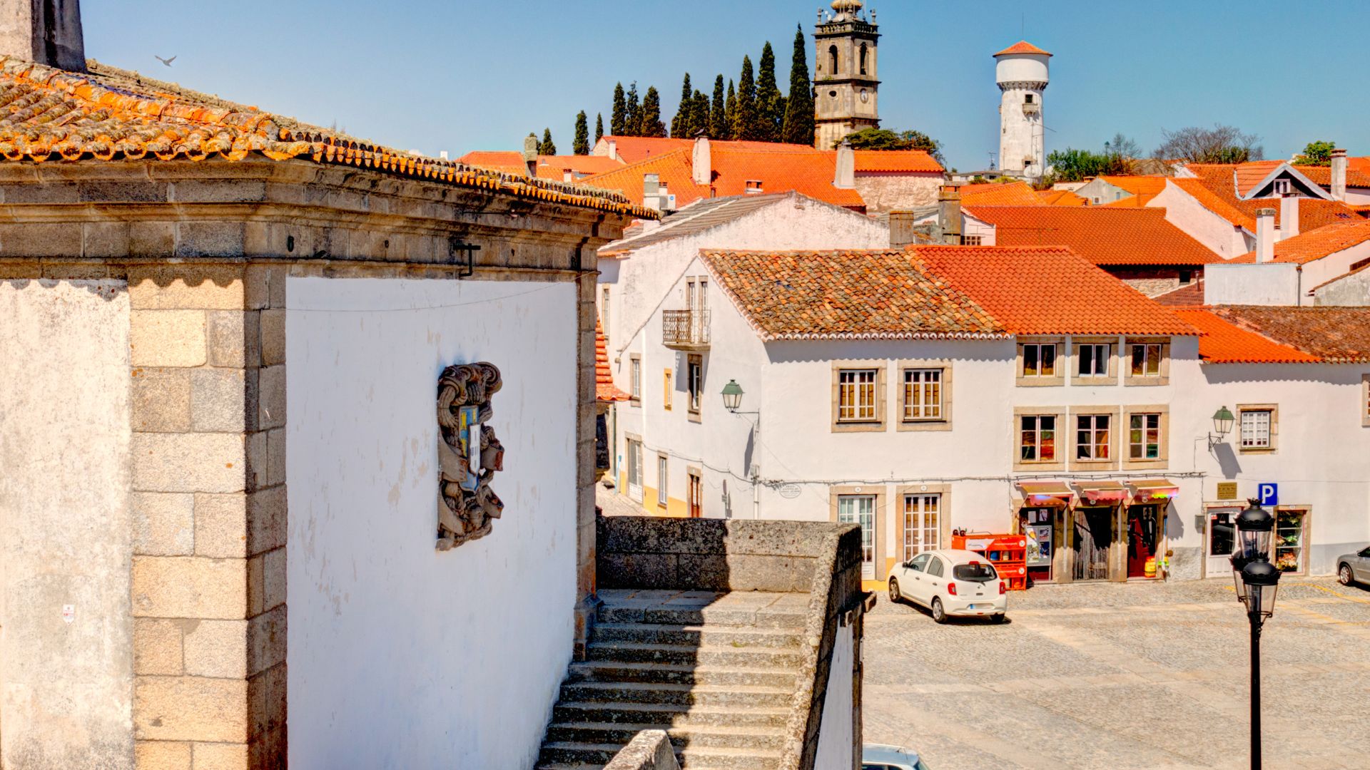 A sunny, elevated view of Almeida, Portugal, showcasing whitewashed buildings with terracotta roofs within the fortified town, a prominent church tower, and a water tower in the distance, all under a clear blue sky.