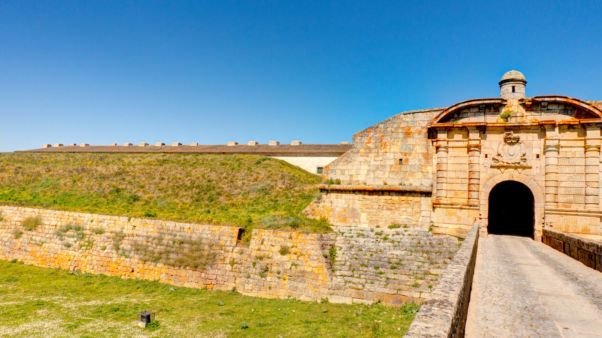 A wide shot of the fortified entrance to Almeida, Portugal, featuring a stone bridge leading to a grand arched gate within a historic star-shaped fortress, with grassy embankments and a clear blue sky above.