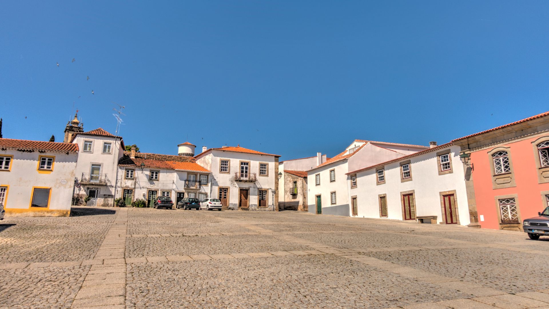 A wide-angle view of a cobblestone town square in Almeida, Portugal, surrounded by traditional white buildings with terracotta roofs under a clear blue sky. The square is empty, and some cars are visible parked along the edges of the buildings.