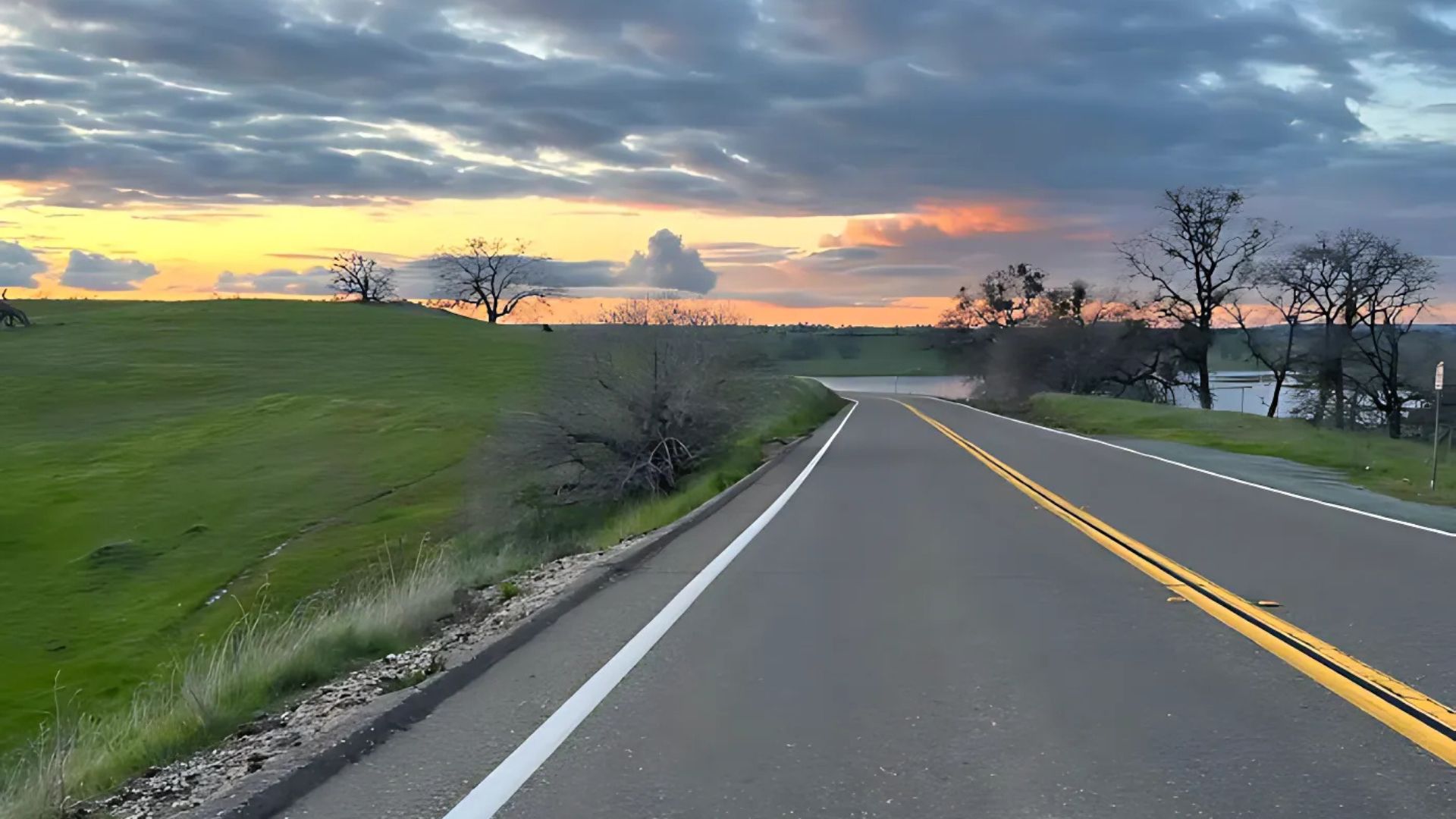 A wide-angle shot of a two-lane asphalt road with a double yellow line on the right and a solid white line on the left, curving gently to the right towards a body of water in the distance. Lush green hills are on the left, and scattered trees, some bare and some with foliage, line the right side of the road and the distant shoreline. The sky above features a dramatic sunset with warm orange and pink hues peeking through a cloudy, grey sky.