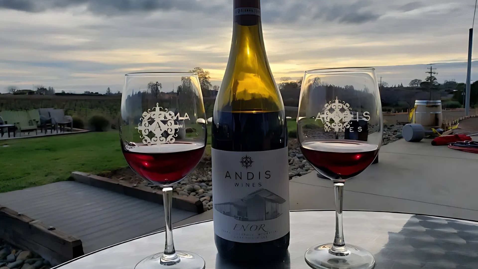A bottle of Andis ENOR red wine stands between two wine glasses filled with red wine on an outdoor table, with a vineyard and cloudy sky in the background at sunset.
