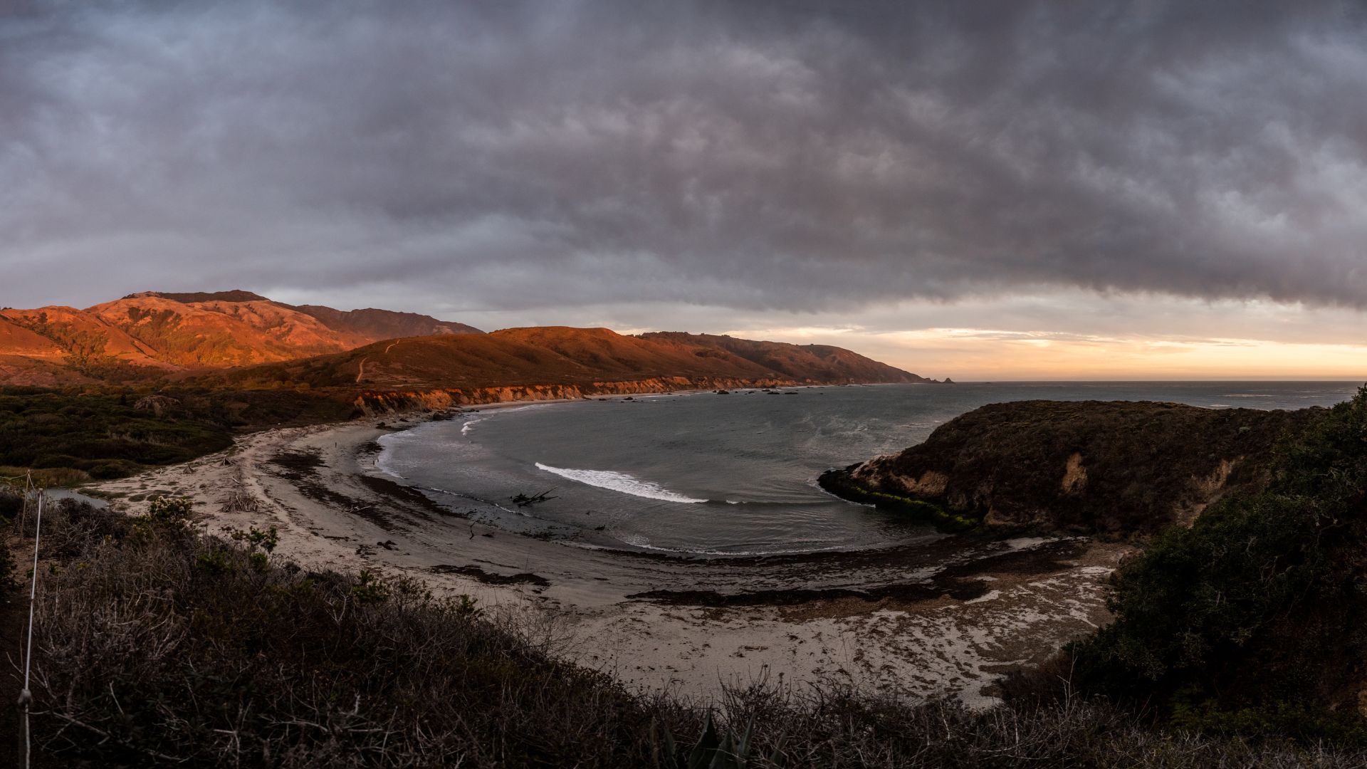 A scenic view of Bixby Creek Bridge, a reinforced concrete open-spandrel arch bridge, spanning a deep canyon along the California coastline, with lush green hills and cliffs meeting the Pacific Ocean on a partly cloudy day. 