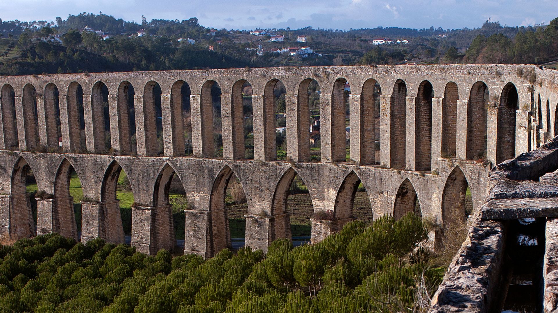 A monumental stone aqueduct with multiple tiers of arches stretching across a valley, surrounded by green trees and distant hills under a clear sky.