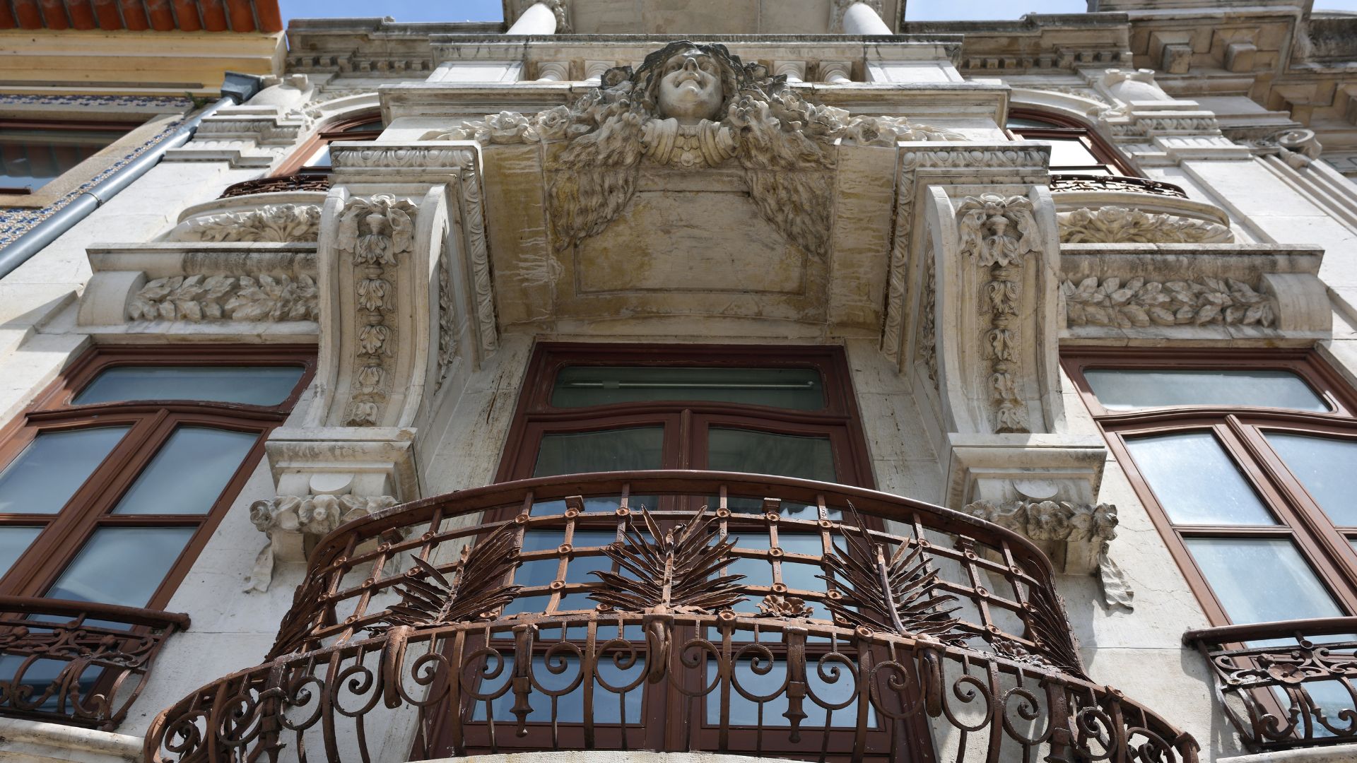 A low-angle shot of an ornate Art Nouveau building facade in Aveiro, Portugal, featuring a detailed stone balcony with a wrought-iron railing and a sculpted female figure above the central window.