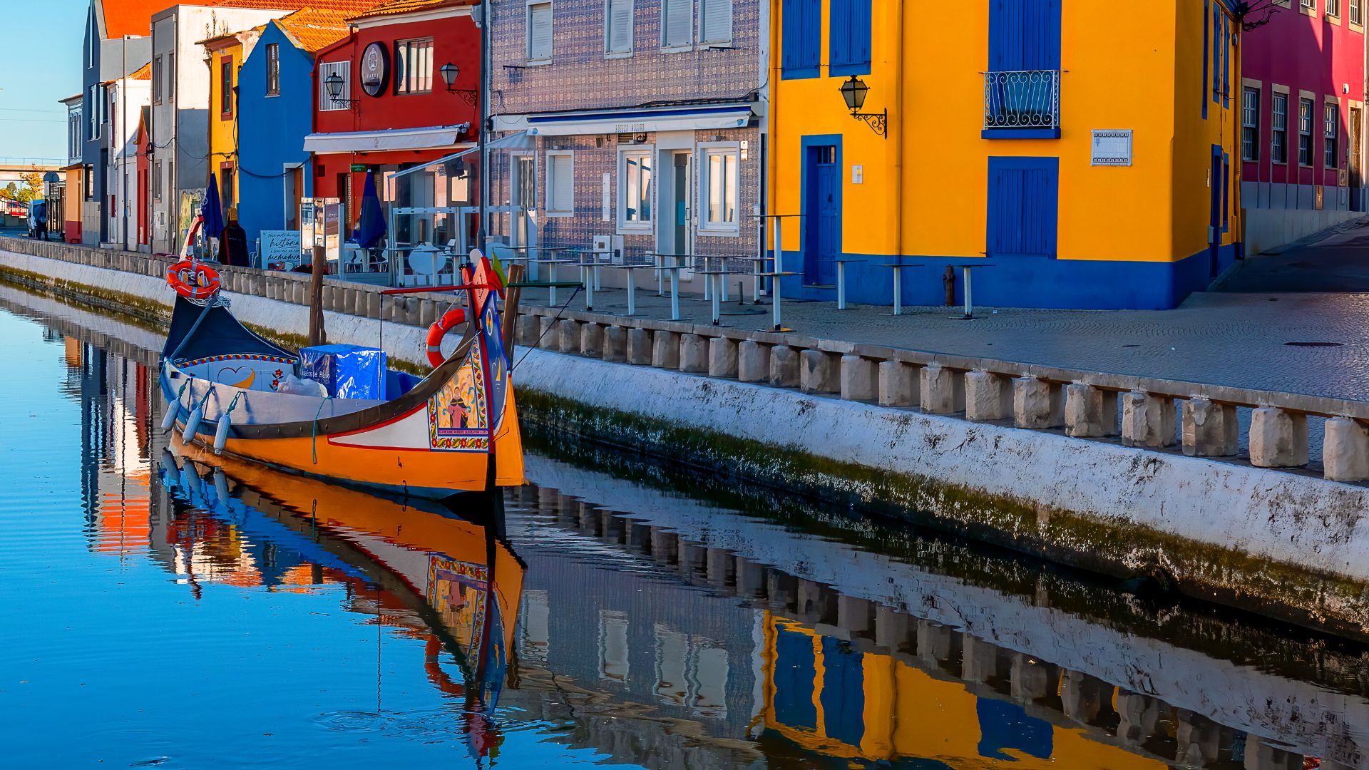 The image captures the picturesque canals of Aveiro, often referred to as the "Venice of Portugal," featuring colorful "moliceiro" boats reflecting on the water, set against a backdrop of charming buildings under a bright blue sky.