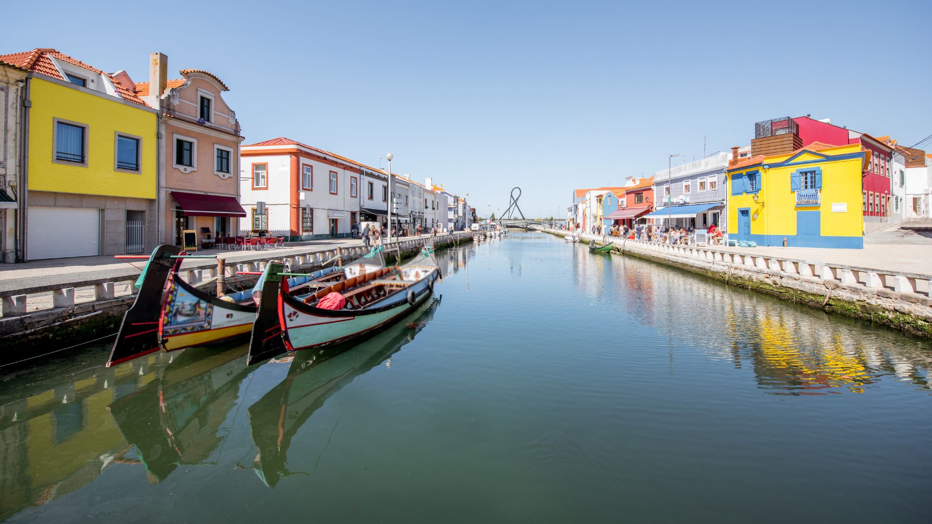 The image captures the picturesque canals of Aveiro, often referred to as the "Venice of Portugal," featuring colorful "moliceiro" boats reflecting on the water, set against a backdrop of charming buildings under a bright blue sky.