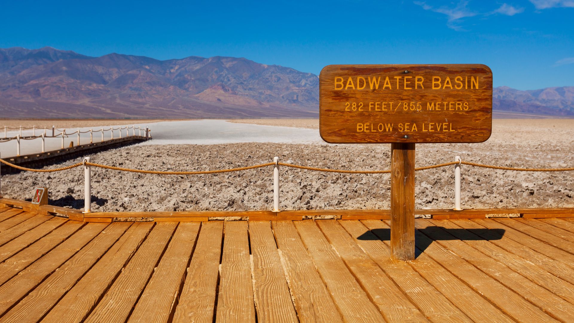 A wooden sign in the foreground reads "BADWATER BASIN 282 FEET/855 METERS BELOW SEA LEVEL," standing on a wooden boardwalk that extends towards the vast, white salt flats of Badwater Basin, with mountains visible in the distance under a clear blue sky.