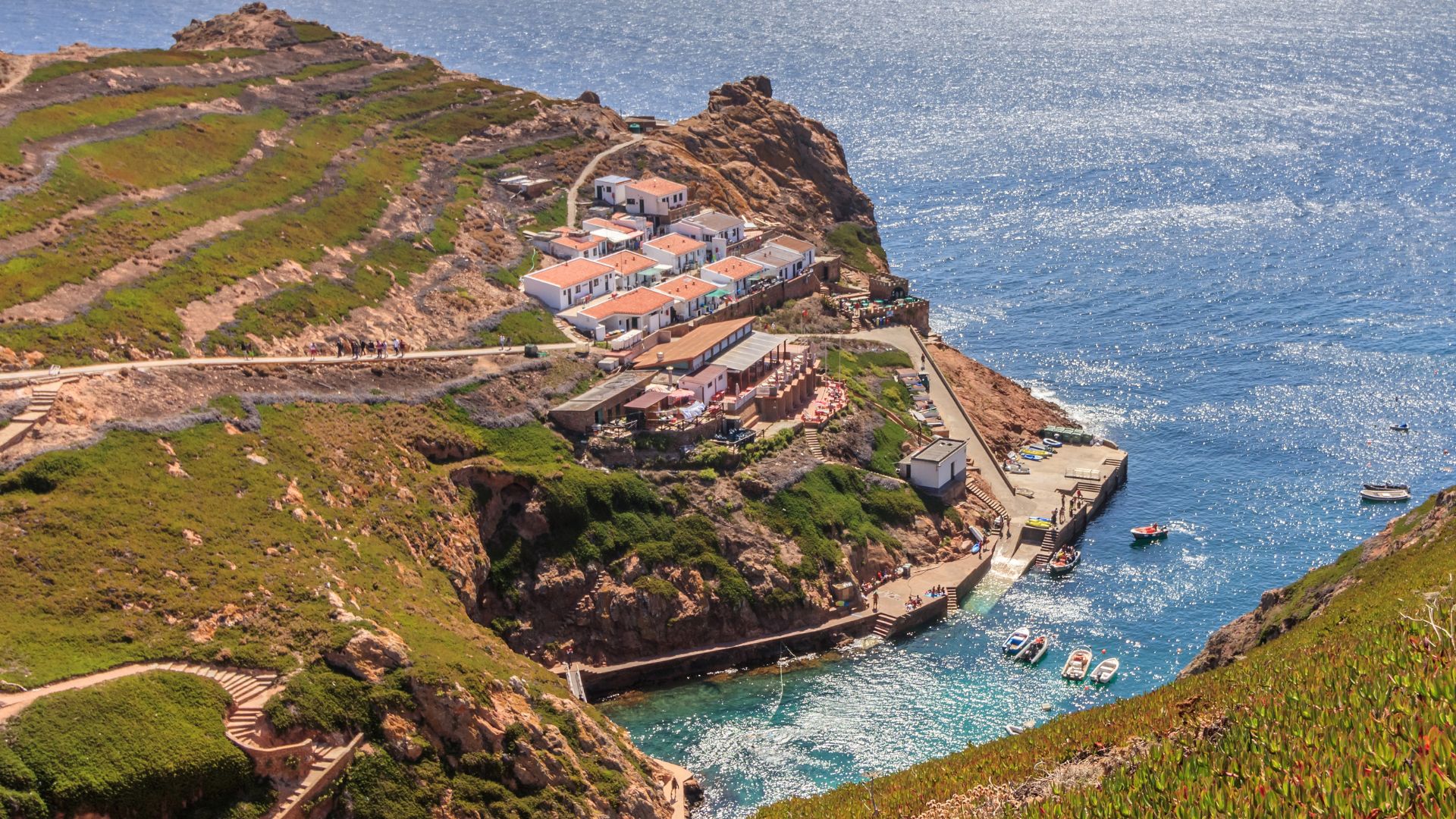The image captures an aerial view of the Berlengas Archipelago, showcasing a cluster of white and red-roofed buildings of Bairro dos Pescadores nestled on the rocky, green cliffs of Berlenga Grande Island. Below the settlement, a small harbor and dock are visible, with boats moored in the clear, blue-green waters of the Atlantic Ocean. The rugged coastline and the vast expanse of the sea frame the scene, with a winding path visible leading down to the harbor. 