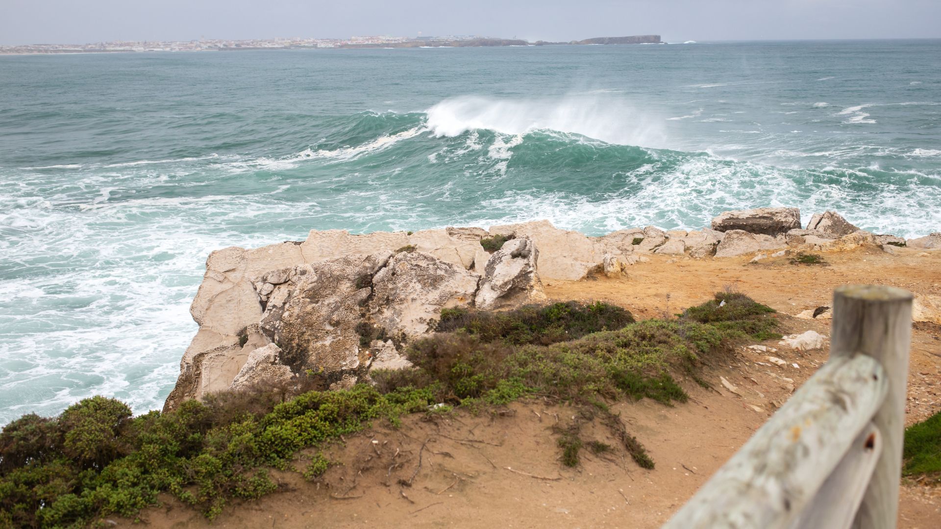 A wide-angle shot of a rugged coastline at Baleal Island, Portugal, featuring large waves crashing against rocky cliffs and a sandy foreground with sparse vegetation, under an overcast sky.