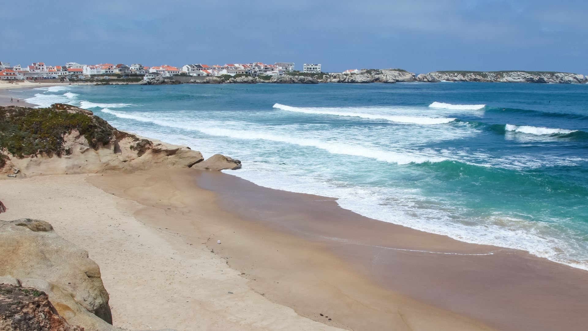 A wide-angle view of Baleal Beach in Peniche, Portugal, featuring a sandy shoreline with breaking waves, a rocky coastline, and a town with white buildings in the distance under a partly cloudy sky.
