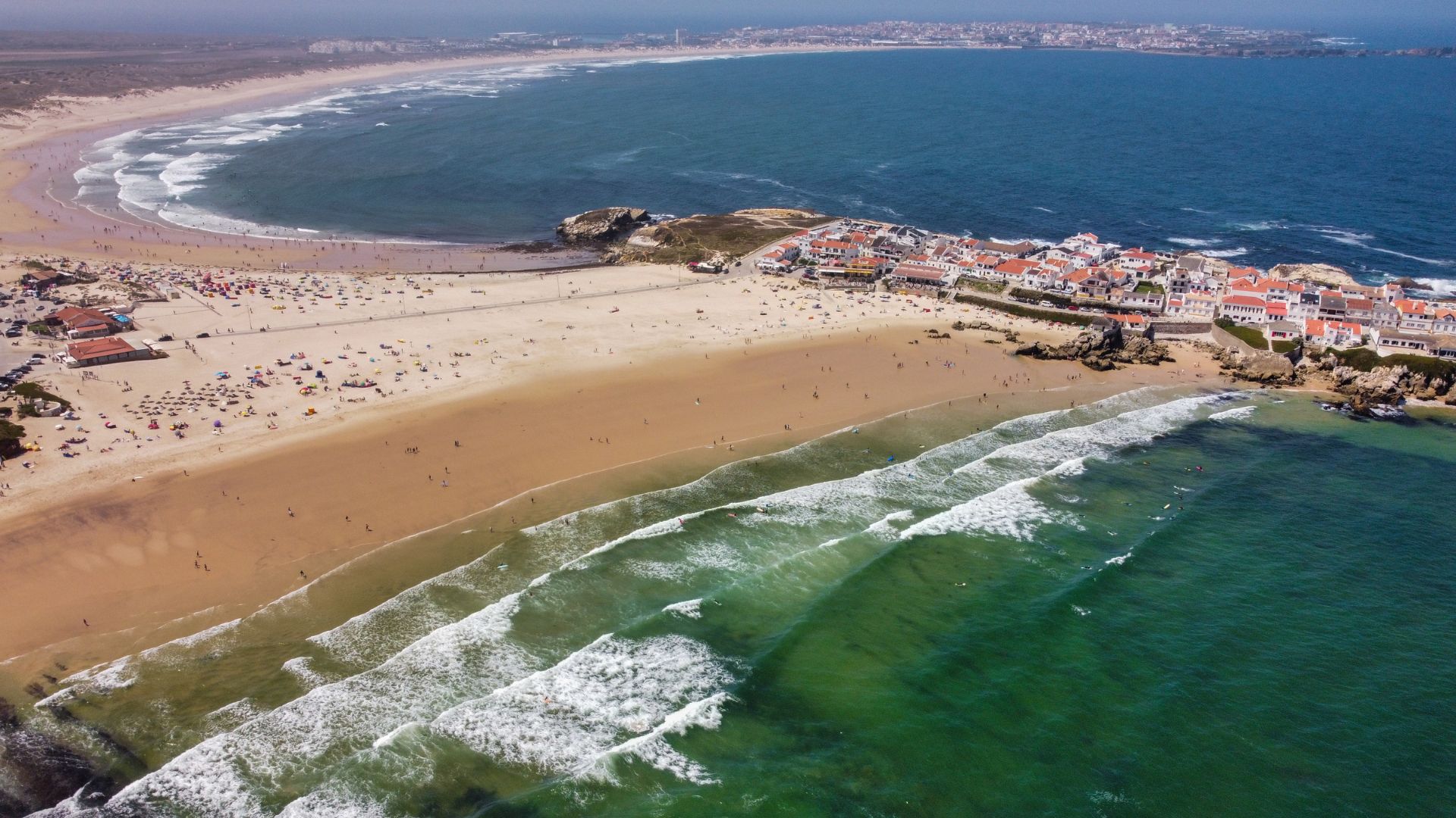 An aerial view of Baleal Beach in Peniche, Portugal, showcasing a sandy peninsula connecting a small island to the mainland, with waves crashing on both sides and a village visible on the island.