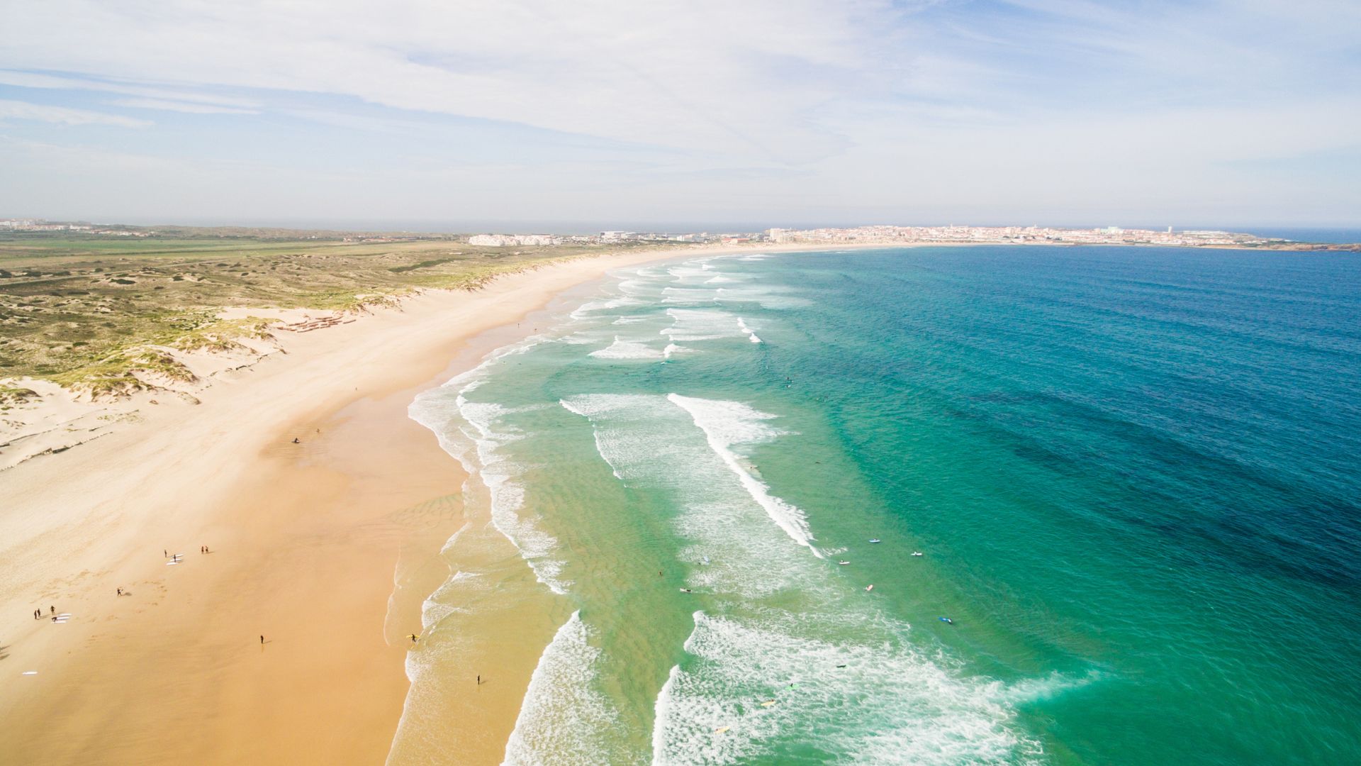 An aerial view of a wide sandy beach with waves breaking on the shore and a small island connected to the mainland by a tombolo, under a bright sky.