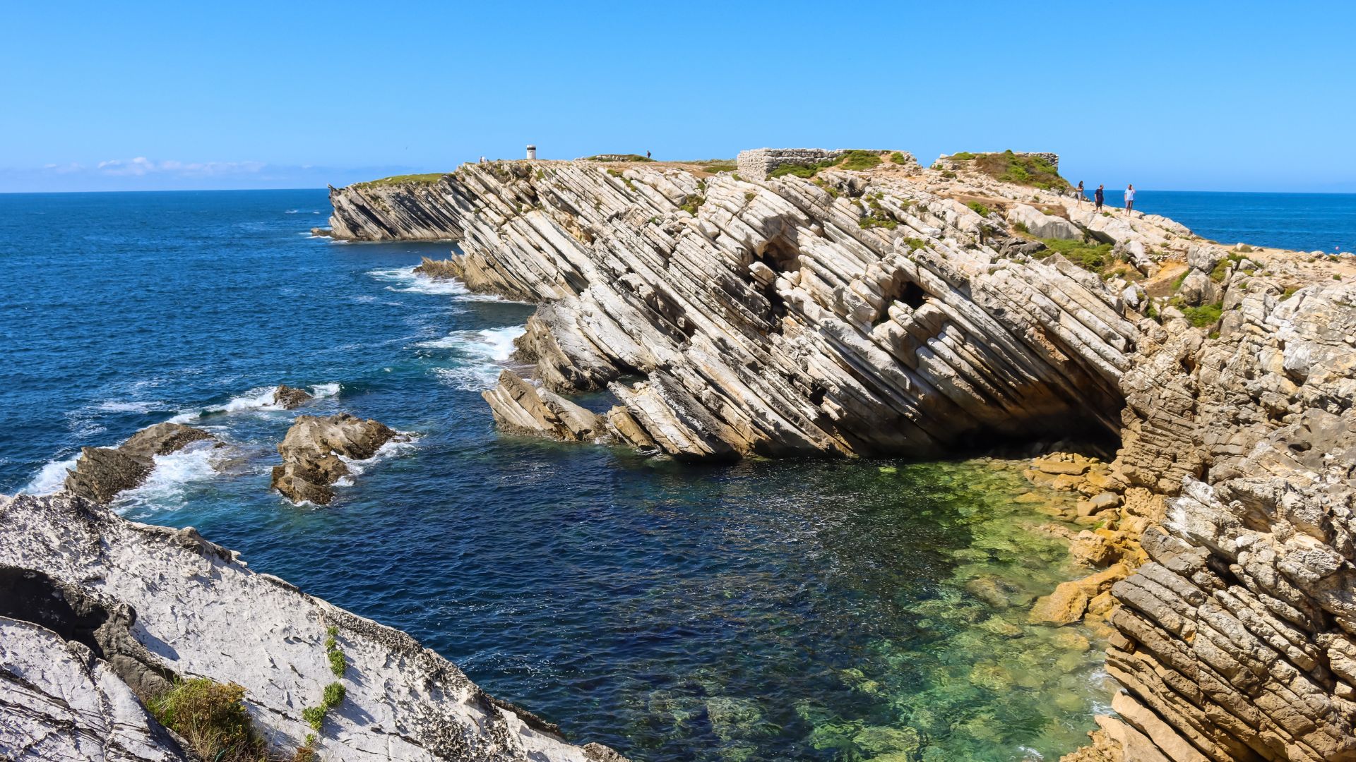 A panoramic view of Baleal Island in Peniche, Portugal, showcasing its distinctive rock formations and cliffs meeting the clear blue waters of the Atlantic Ocean under a bright sky. The island is connected to the mainland by sandy beaches, and the water near the shore reveals a vibrant green hue.