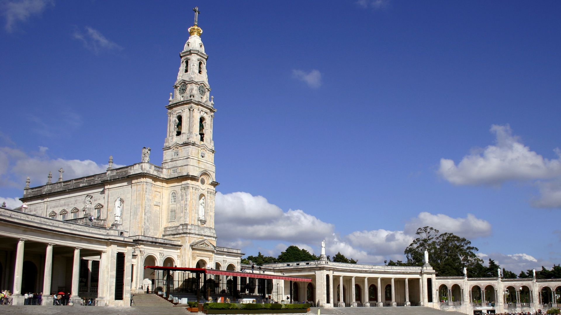 A wide shot of the Sanctuary of Our Lady of Fátima in central Portugal, featuring the prominent white Basilica of Our Lady of the Rosary with its tall bell tower, set against a bright blue sky with scattered white clouds, and surrounded by colonnades on either side. 