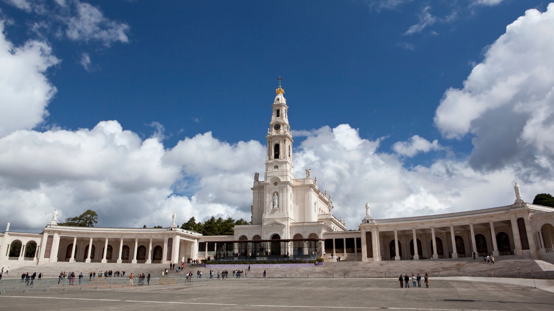 A wide shot of the Sanctuary of Our Lady of Fátima in central Portugal, featuring the prominent white Basilica of Our Lady of the Rosary with its tall bell tower under a bright blue sky with scattered clouds. The foreground