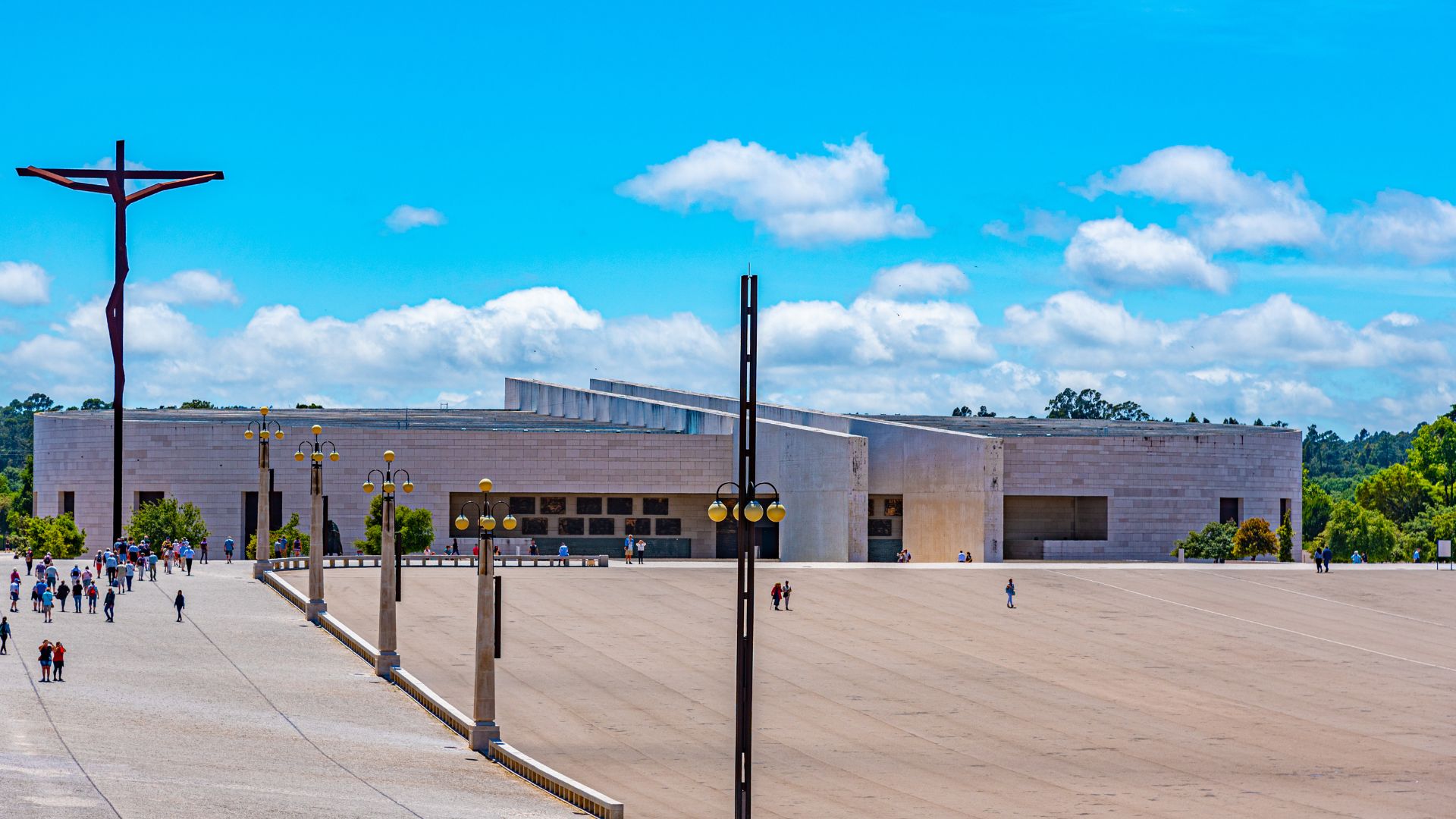 A wide shot of the modern, expansive Basilica of the Holy Trinity in Fátima, Portugal, with a large open plaza leading up to it, and a tall, dark cross monument to the left.