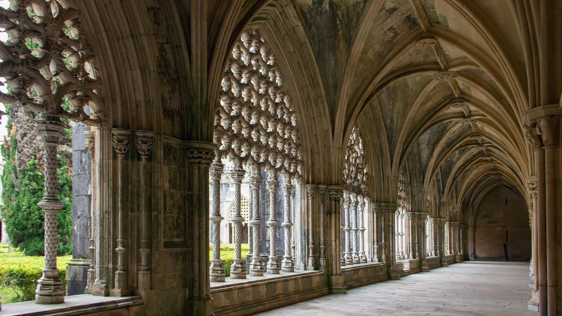 A long, arched stone hallway within a historic monastery, featuring ornate Gothic and Manueline architectural details, including intricate window tracery and vaulted ceilings, leading towards an open courtyard with greenery visible through the windows.