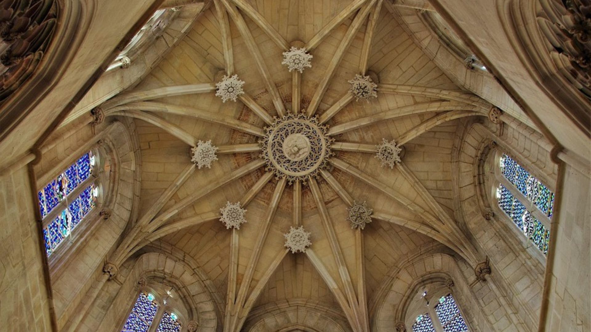 A low-angle interior view of the Batalha Monastery in Portugal, showcasing its intricate Gothic and Manueline vaulted ceiling with radiating ribs and a central boss, surrounded by tall arched windows with colorful stained glass, and illuminated by sunlight streaming through the windows.