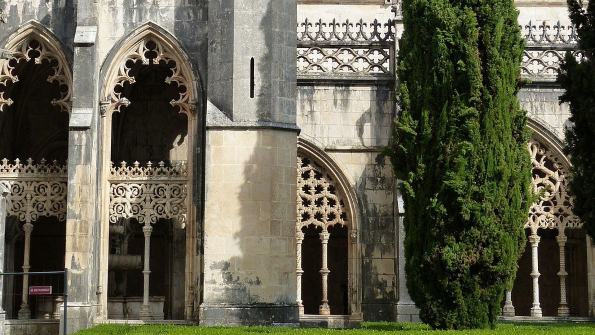 A view of the Batalha Monastery in Portugal, showcasing its intricate Gothic architecture, including pointed arches, ornate stonework, and a tall central spire, with a slender cypress tree standing prominently in the foreground against a partly cloudy sky.