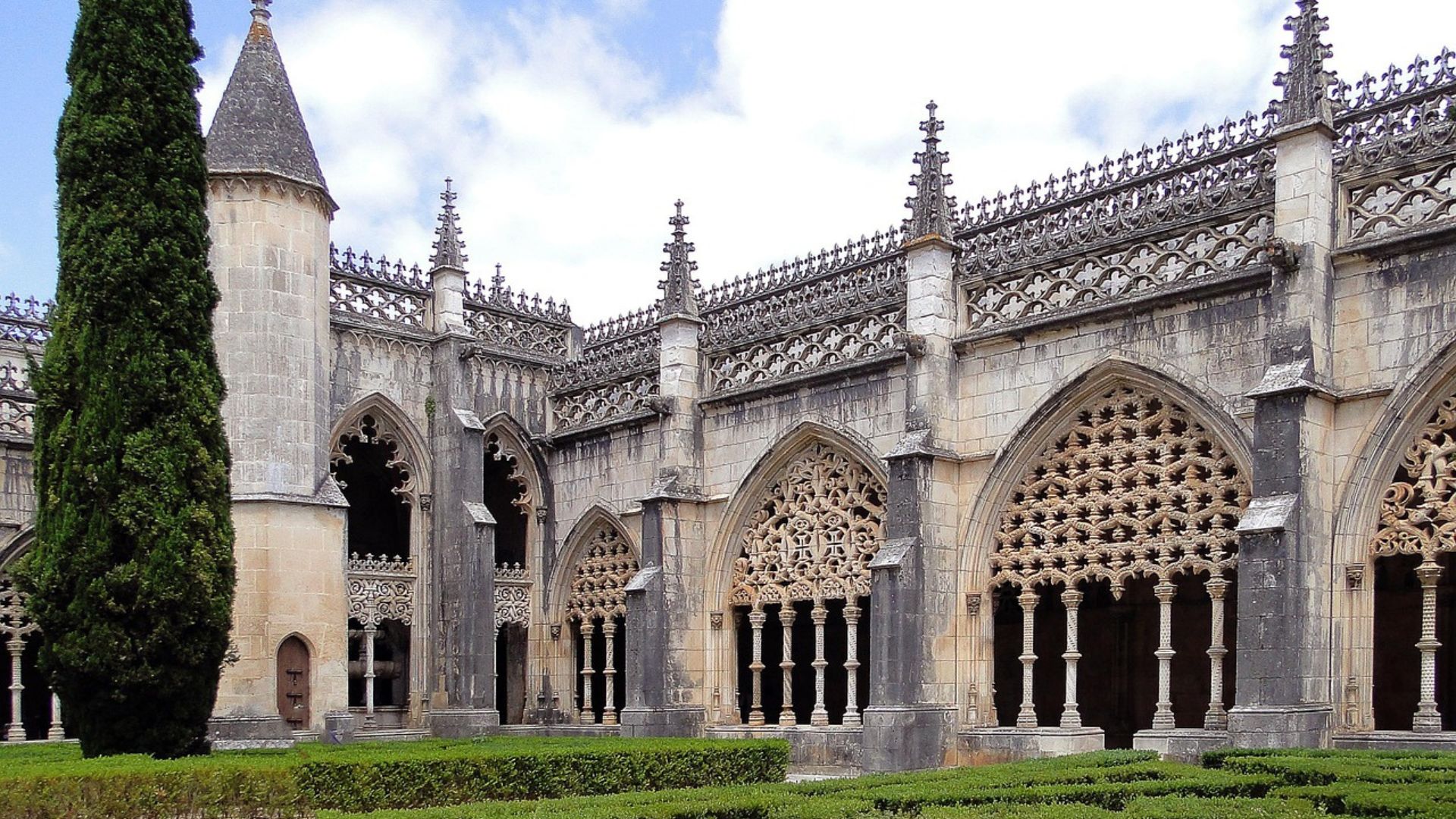 A grand, ornate Gothic monastery with intricate stonework and arched windows, surrounded by a manicured green garden with hedges, under a partly cloudy sky.