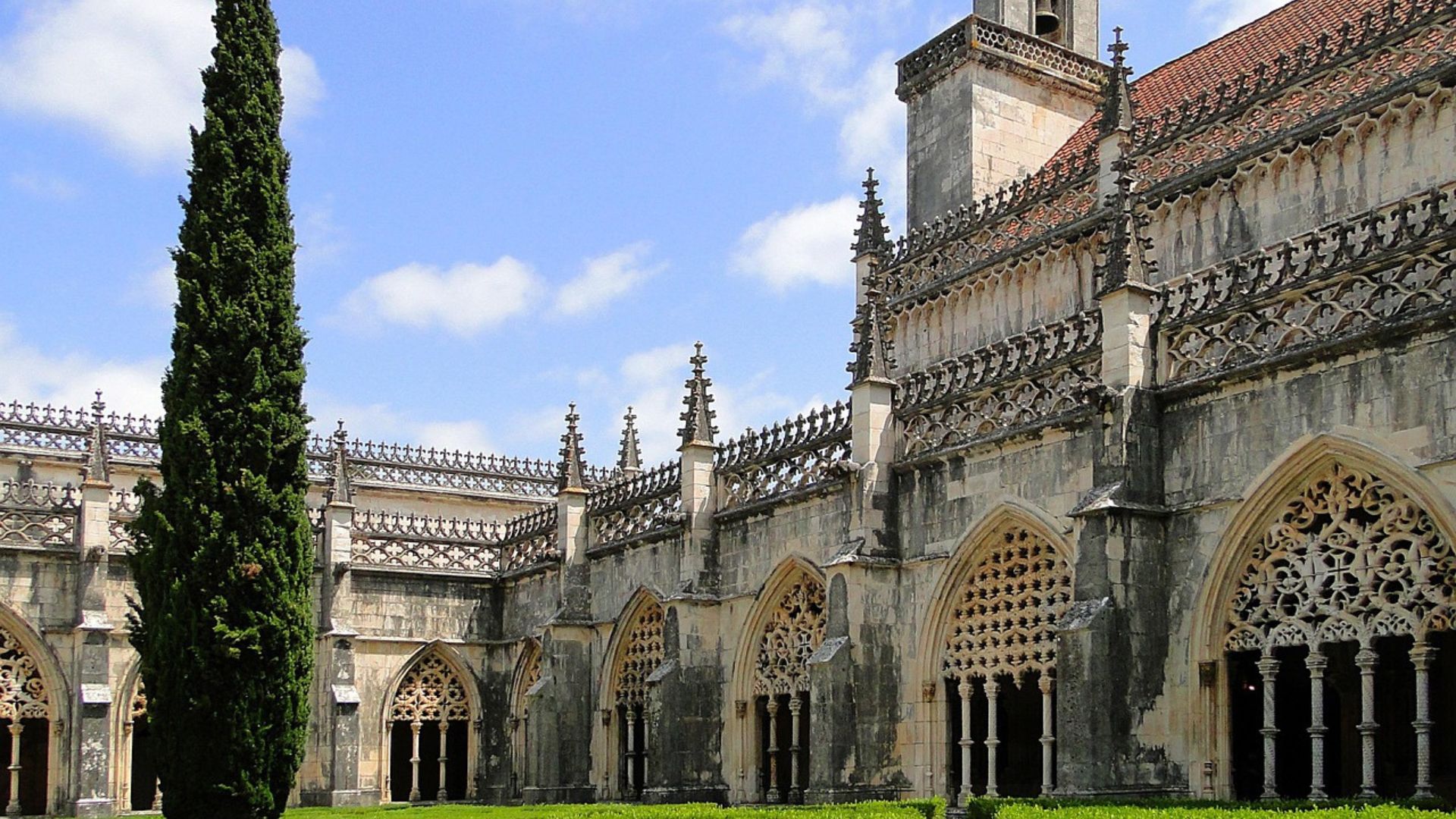 A grand, ornate Gothic monastery with intricate stonework and arched windows, surrounded by a manicured green garden with hedges, under a partly cloudy sky.