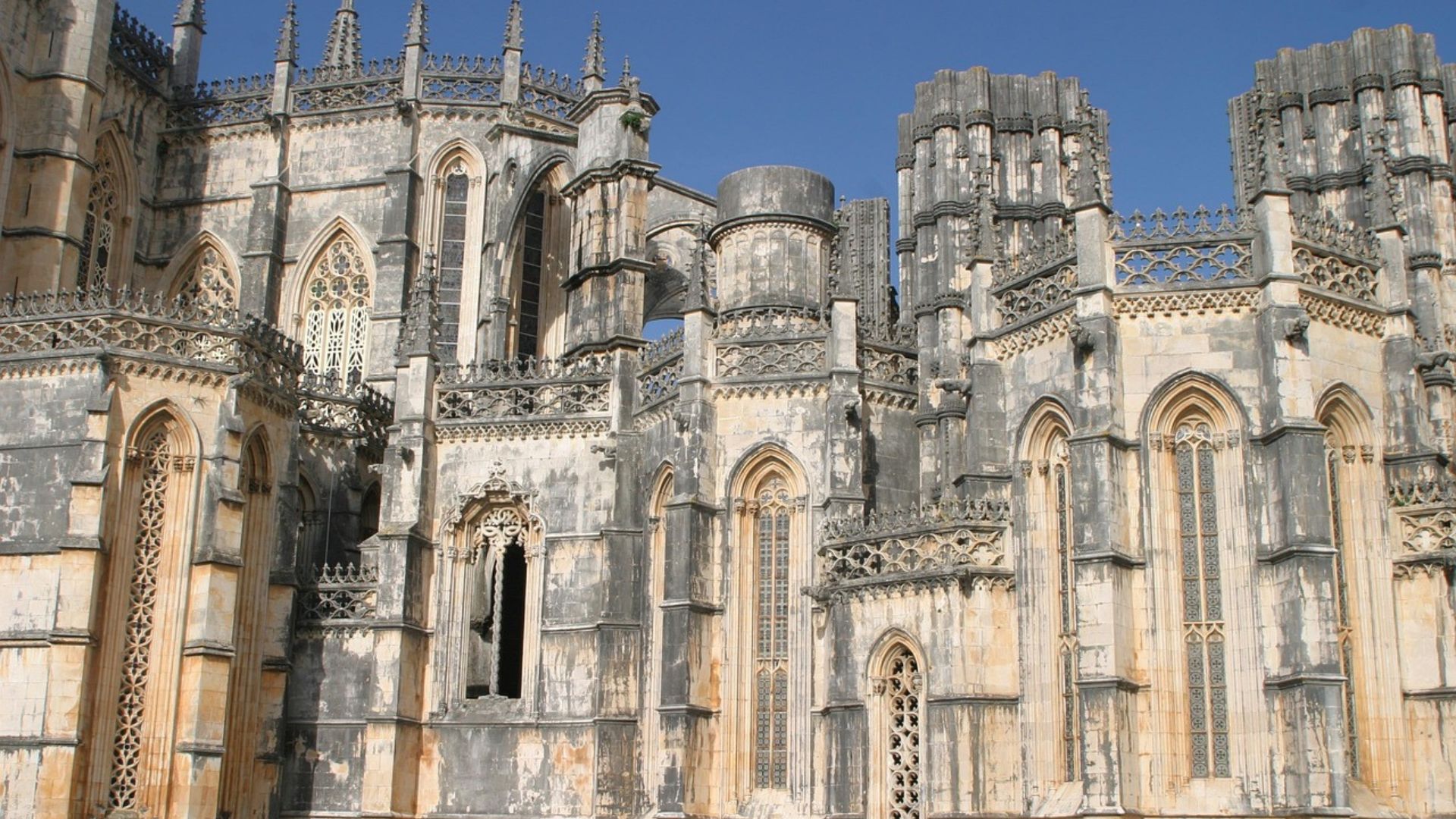 A detailed, low-angle shot of the Batalha Monastery in Portugal, showcasing its intricate Gothic and Manueline architectural details, including ornate stonework, spires, and arched windows, set against a clear blue sky.
