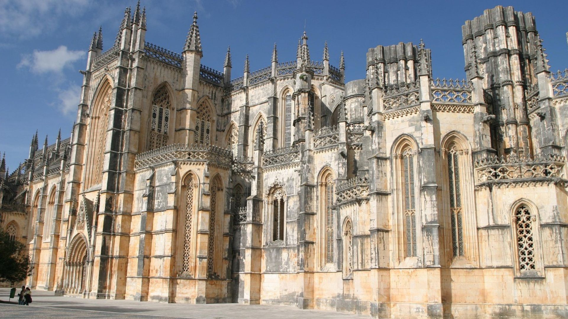A grand, ornate Gothic monastery with numerous spires, pinnacles, and intricate stonework under a clear blue sky, set against a paved foreground.