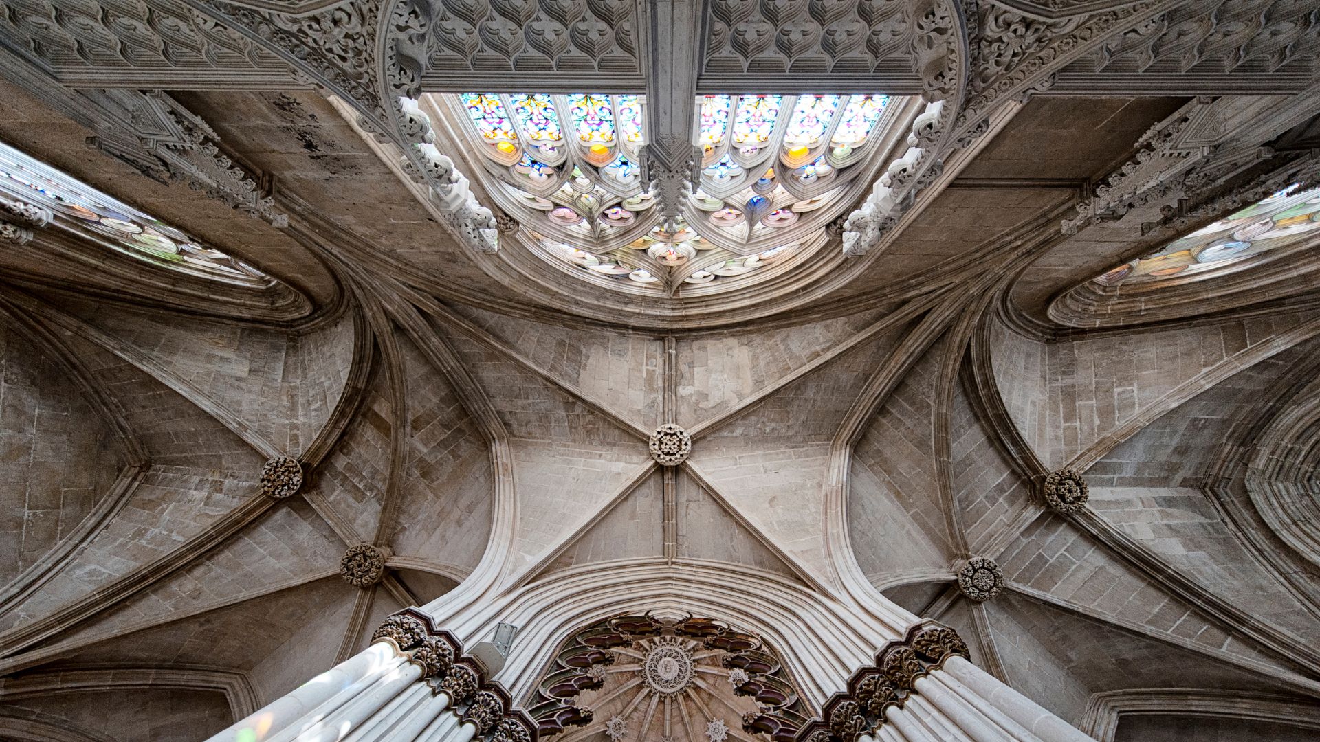 A low-angle view showcasing the elaborately vaulted stone ceiling and stained-glass windows of the Batalha Monastery in Portugal, highlighting its detailed Gothic and Manueline architectural styles. 