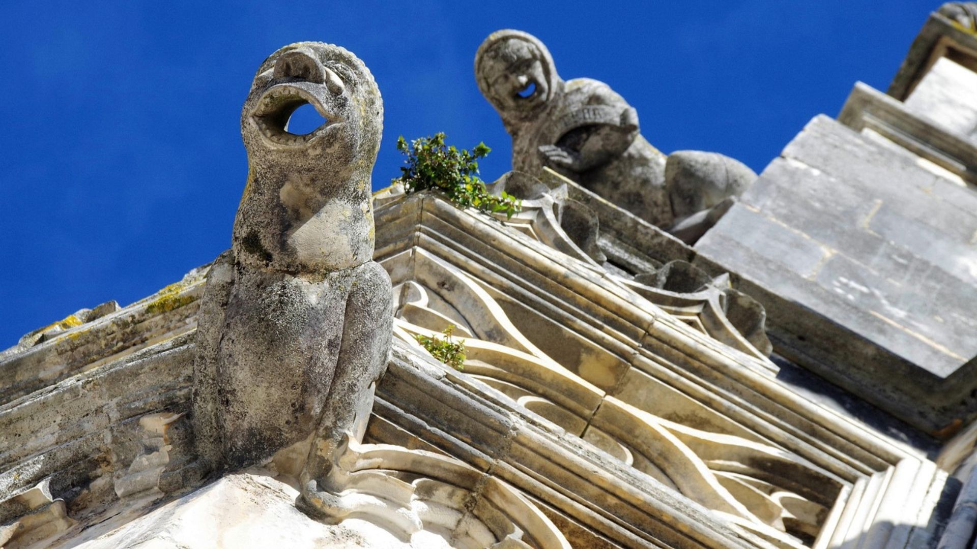 A close-up view of two intricately carved stone gargoyles on the exterior of a Gothic-style building, likely part of the Batalha Monastery, under a clear blue sky.
