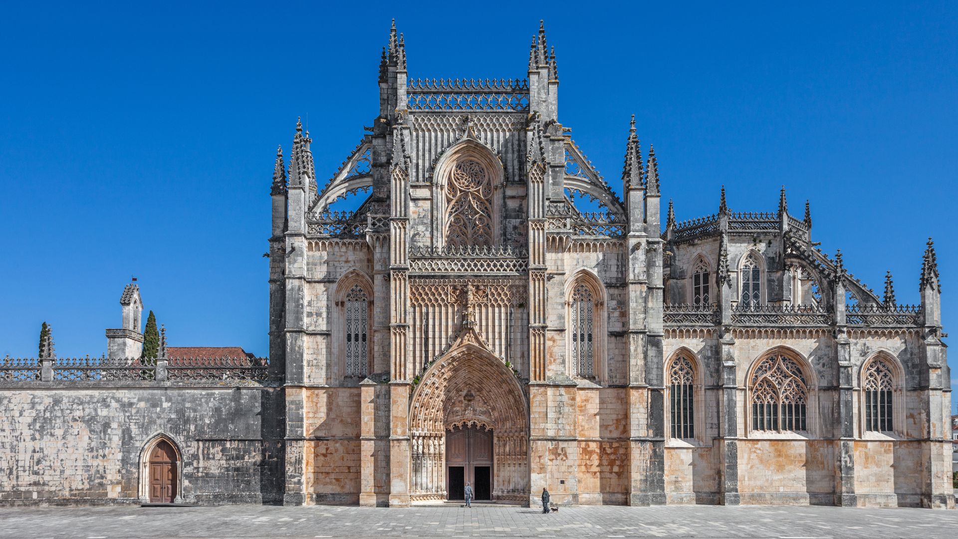A grand, ornate Gothic monastery with intricate stone carvings and tall arched windows, featuring a prominent entrance portal, set against a clear blue sky.
