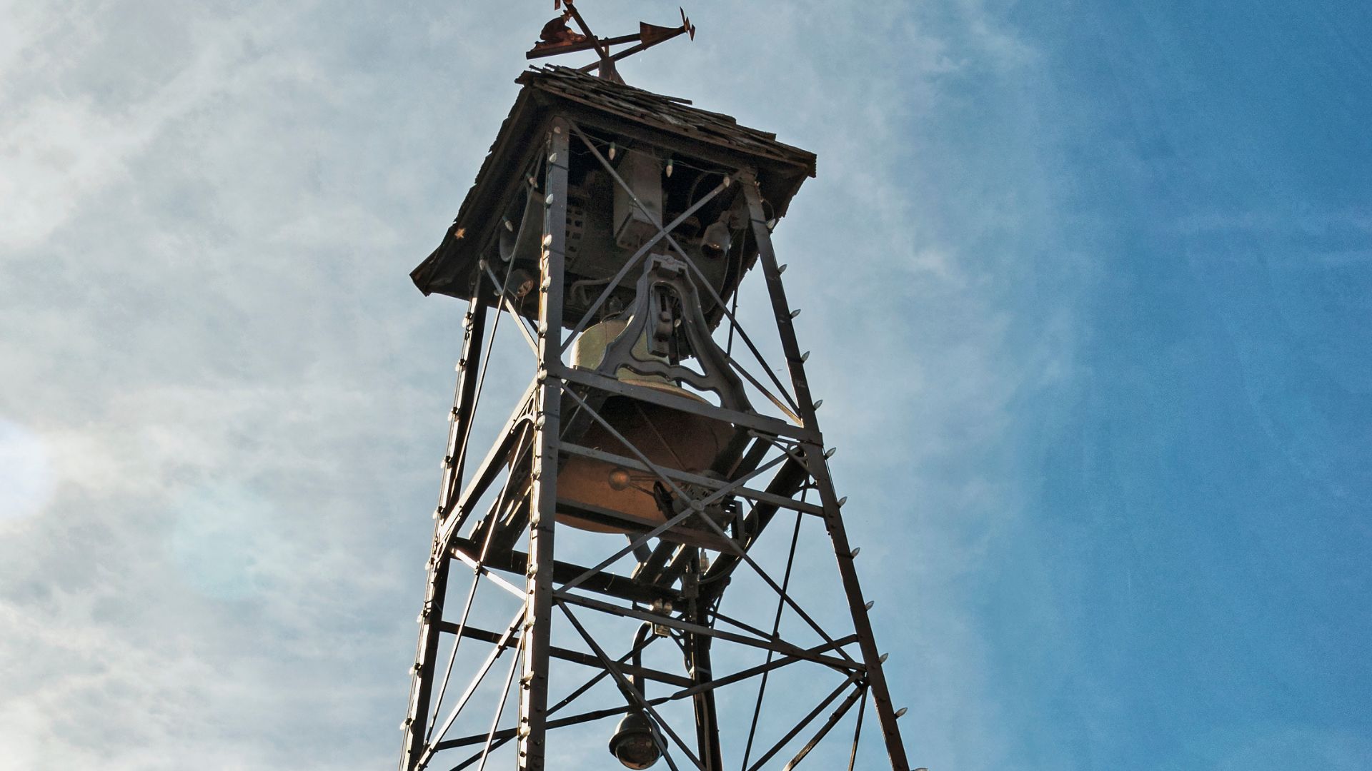 A low-angle shot of the historic Bell Tower in Placerville, California, showing its weathered wooden and metal structure, the bell visible within its open belfry, against a vibrant blue sky.