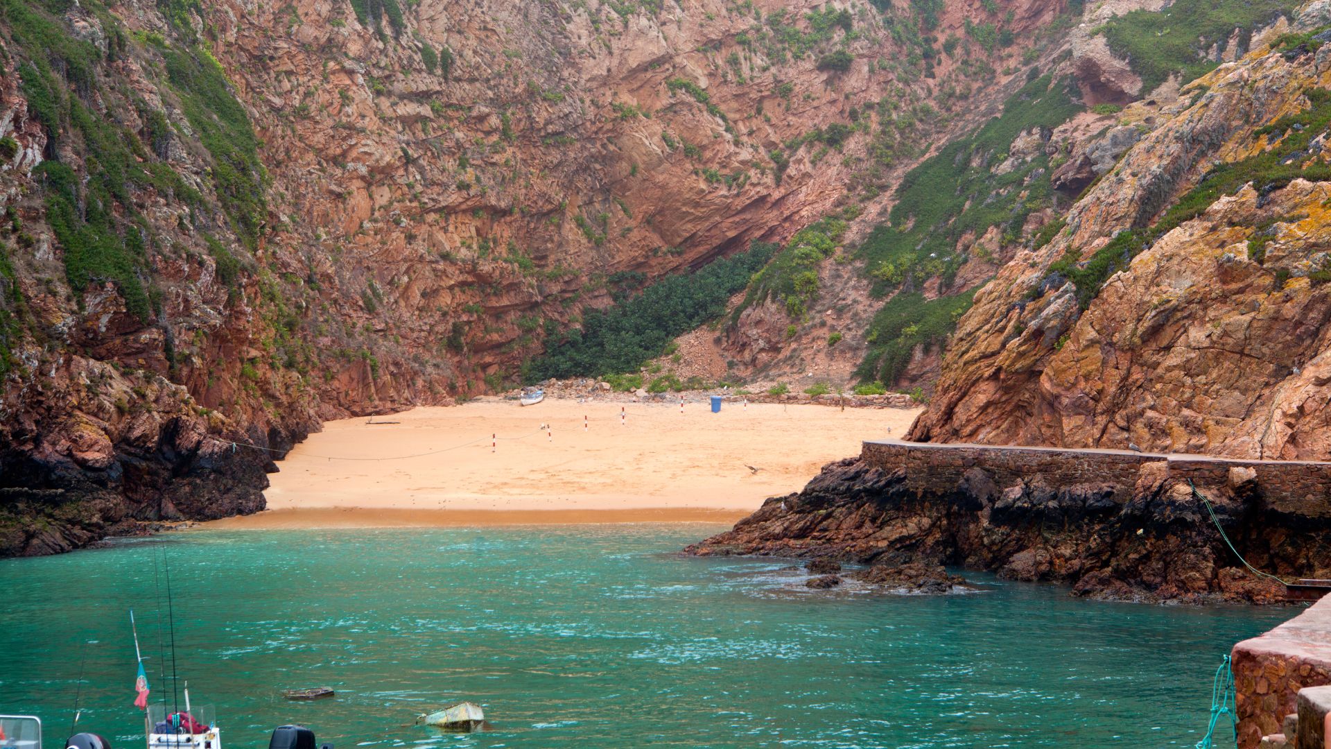 A view of a secluded sandy beach nestled between rugged, rocky cliffs, with clear turquoise water in the foreground and a small pier extending from the right side.