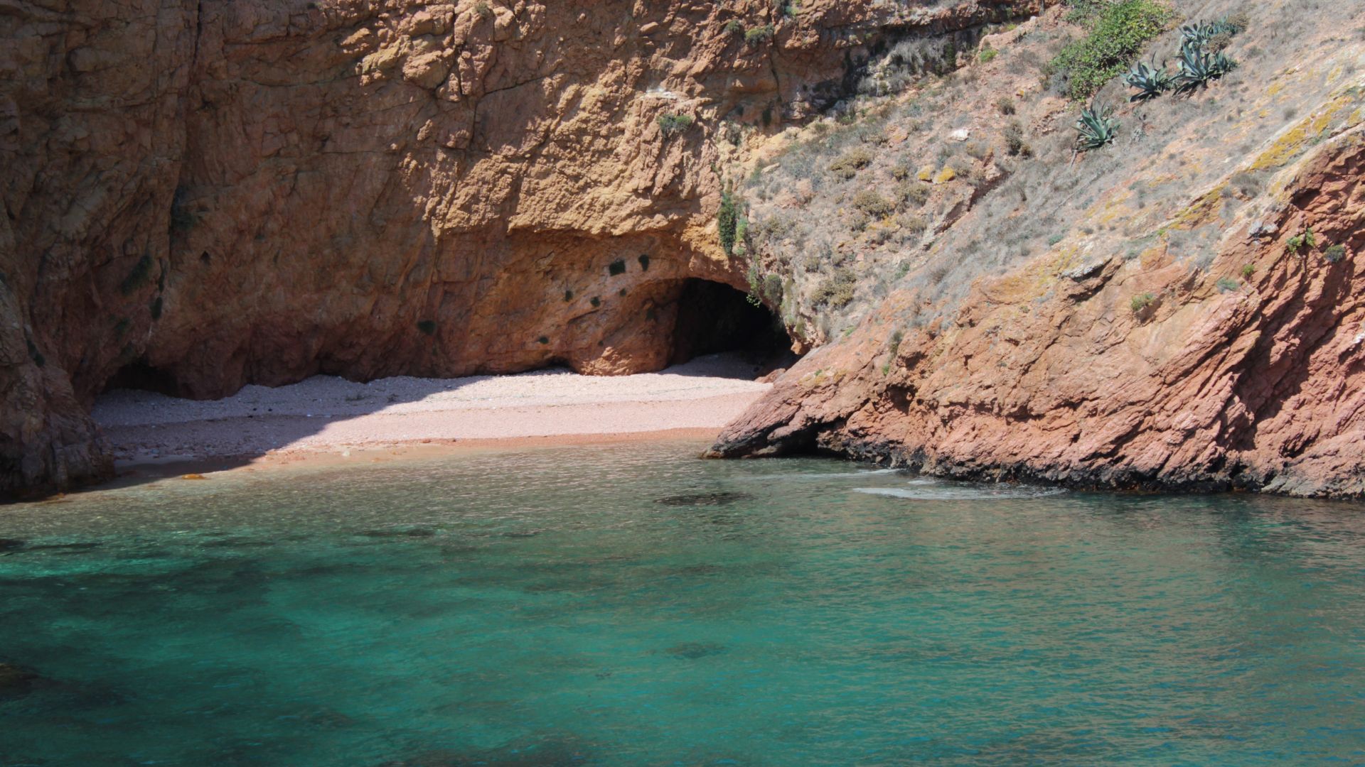 A serene cove with a small sandy beach nestled between rugged cliffs, leading into clear turquoise waters, with a dark cave entrance visible in the cliff face.