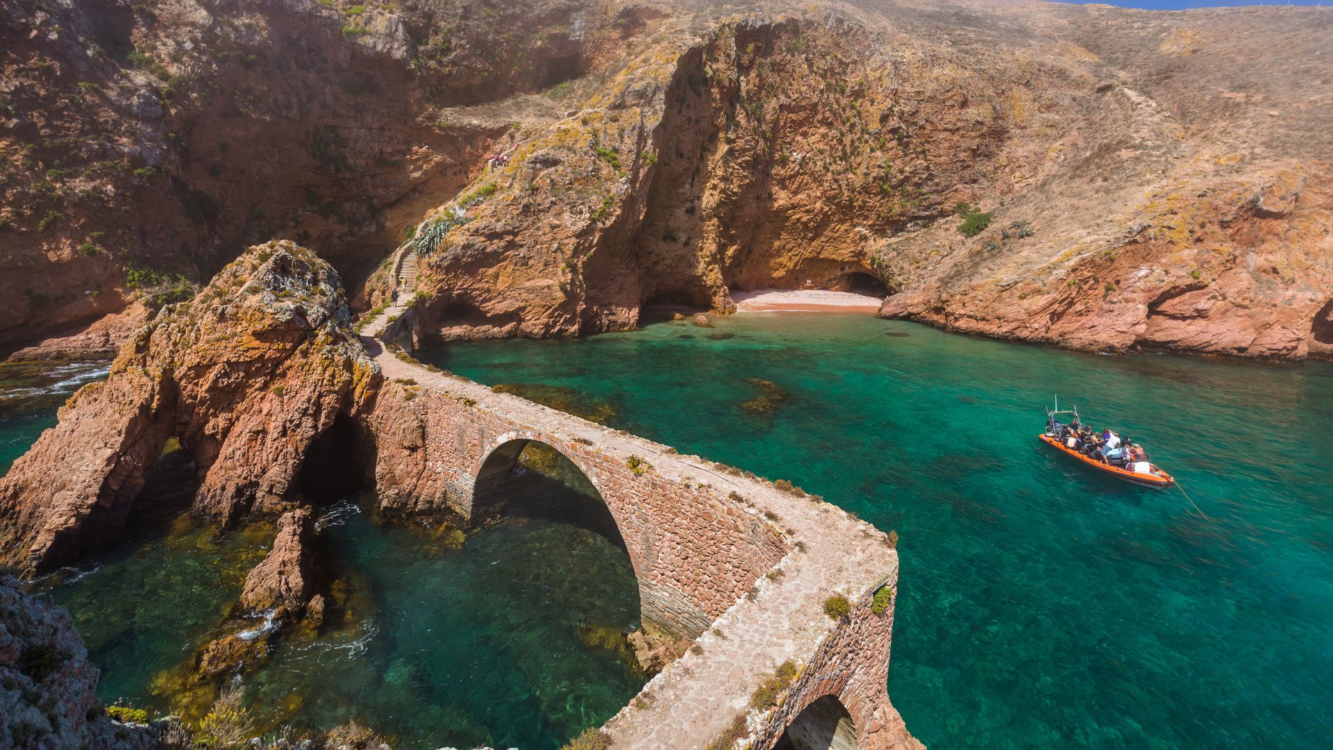 An aerial view of the Fort of São João Baptista, a historic fortress with a stone bridge connecting it to the main island of Berlenga Grande, surrounded by clear turquoise waters and rugged cliffs. A small boat is visible near the fort.