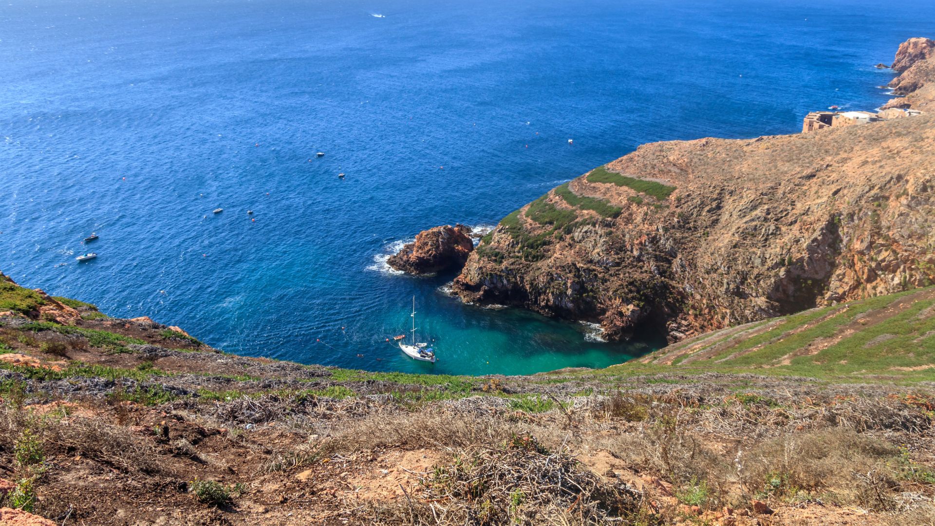 The image depicts a dramatic coastal landscape featuring a deep blue sea with a turquoise cove, surrounded by rugged, rocky cliffs covered in sparse vegetation. A small sailboat is anchored in the clear waters of the cove, while other smaller boats are visible further out in the ocean. The perspective is from a high vantage point, looking down into the cove and out towards the open sea.
