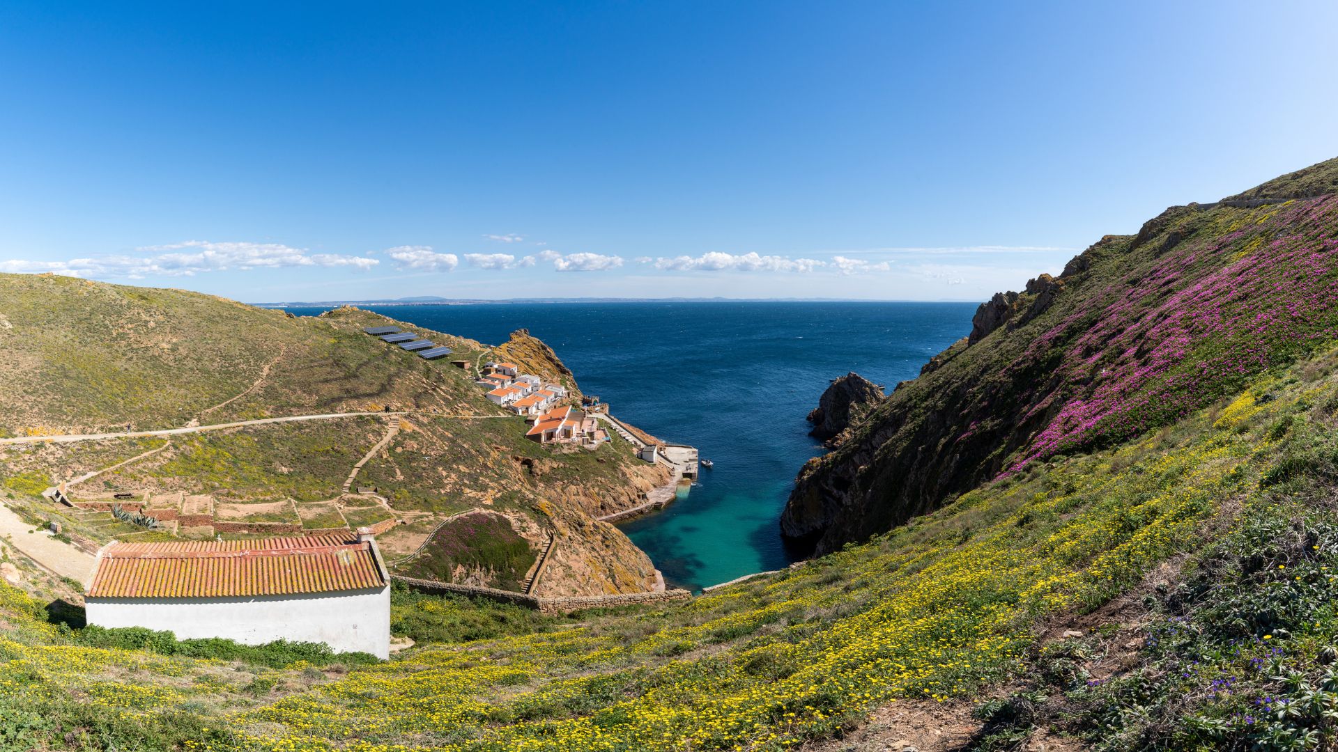 The image depicts a dramatic coastal landscape featuring a deep blue sea with a turquoise cove, surrounded by rugged, rocky cliffs covered in sparse vegetation. A small sailboat is anchored in the clear waters of the cove, while other smaller boats are visible further out in the ocean. The perspective is from a high vantage point, looking down into the cove and out towards the open sea.