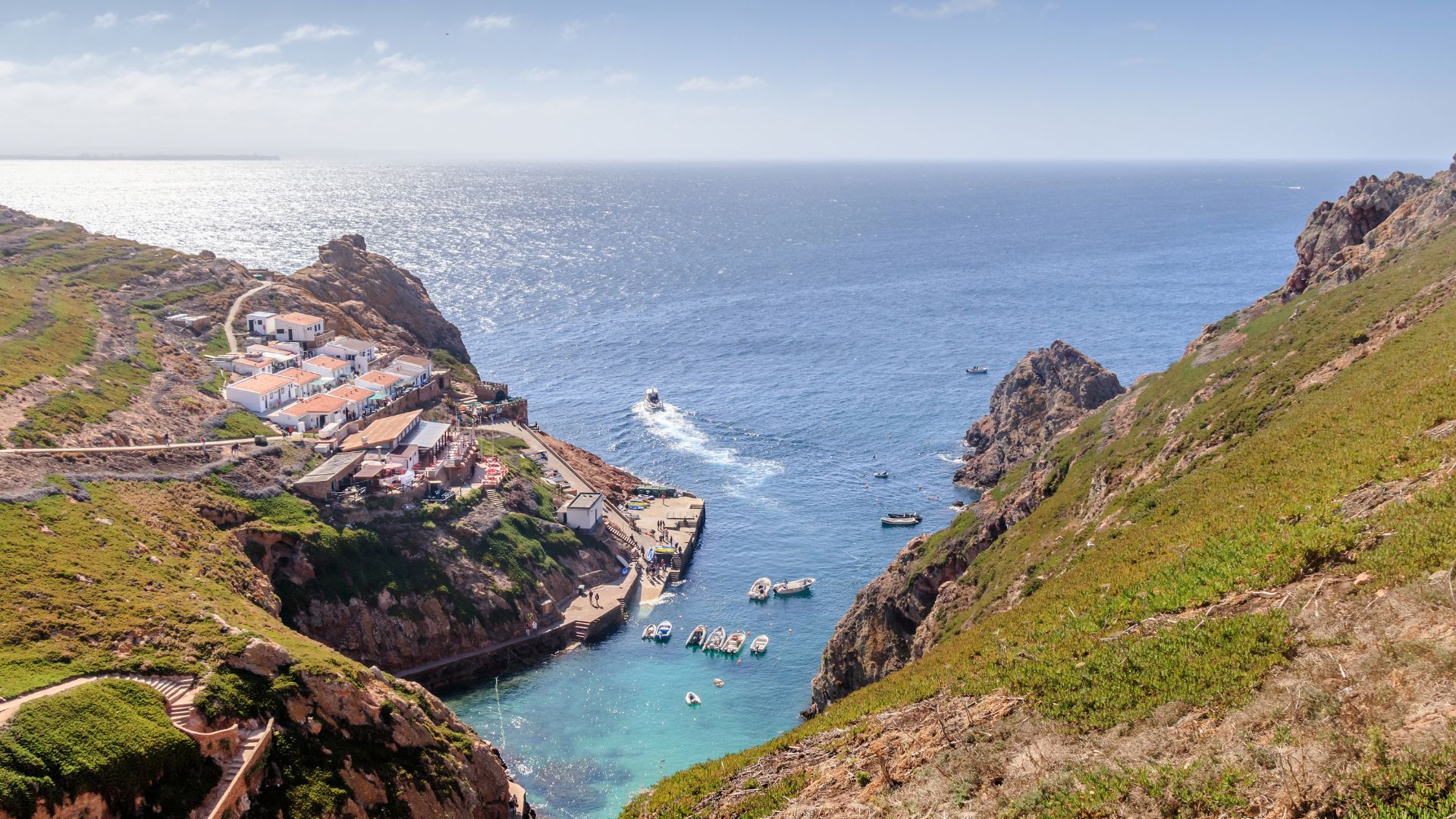 An aerial view capturing the rugged, green cliffs of the Berlengas Archipelago in Portugal, revealing a small village nestled in a cove with boats in the turquoise water, and the vast expanse of the Atlantic Ocean beyond.
