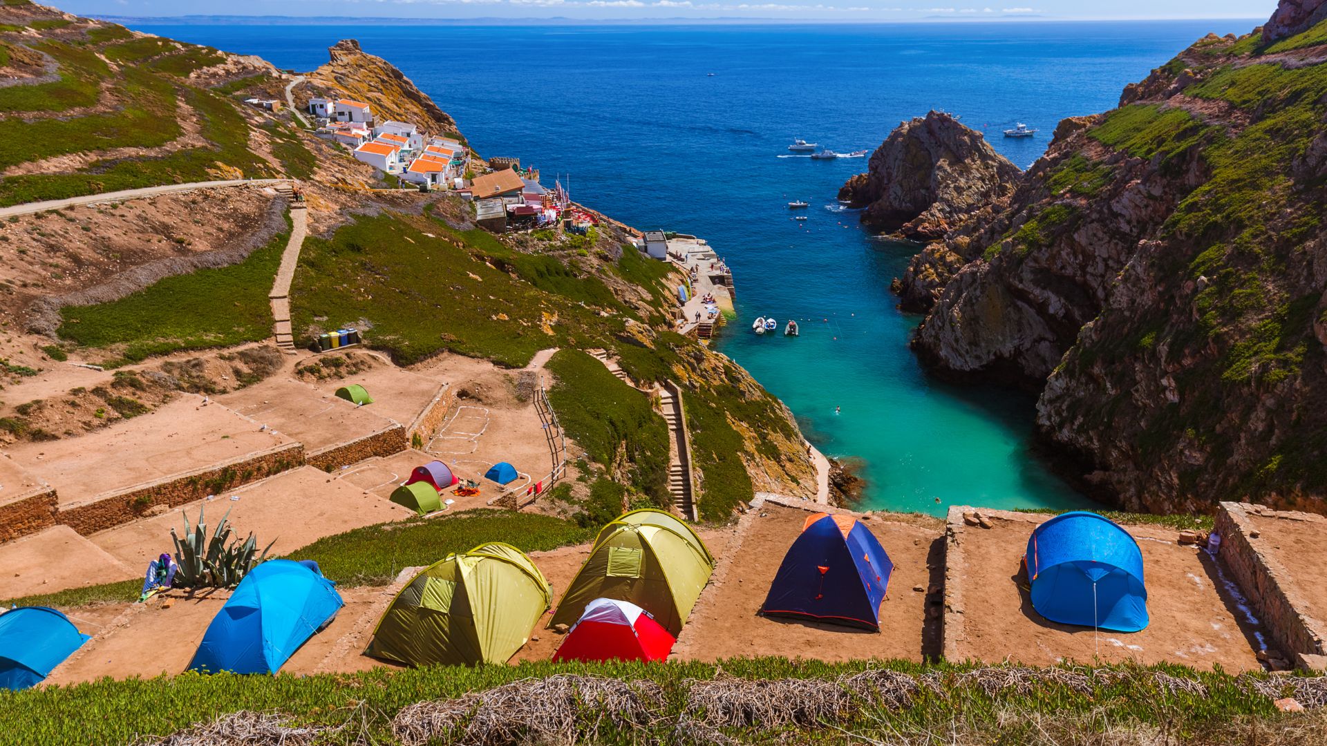 An aerial view captures the scenic Berlengas archipelago in Portugal, featuring a campsite with several colorful tents pitched on terraced ground overlooking a turquoise cove. In the background, white-washed buildings of a small village cling to the hillside above the clear blue sea, with rocky cliffs and boats dotting the water. 