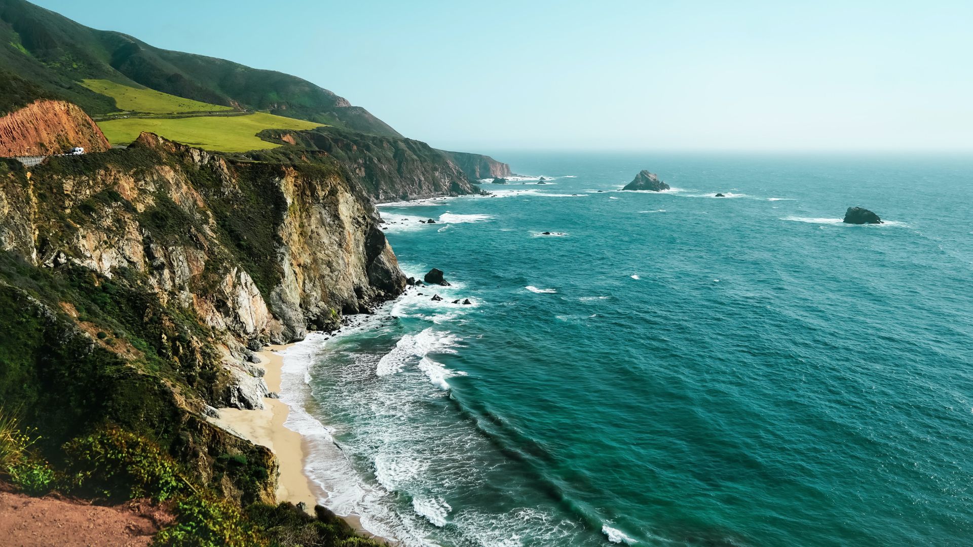 A breathtaking coastal landscape showing the rugged cliffs of Big Sur, California, meeting the turquoise waters of the Pacific Ocean, with waves crashing onto a sandy beach and a winding road visible along the clifftop.
