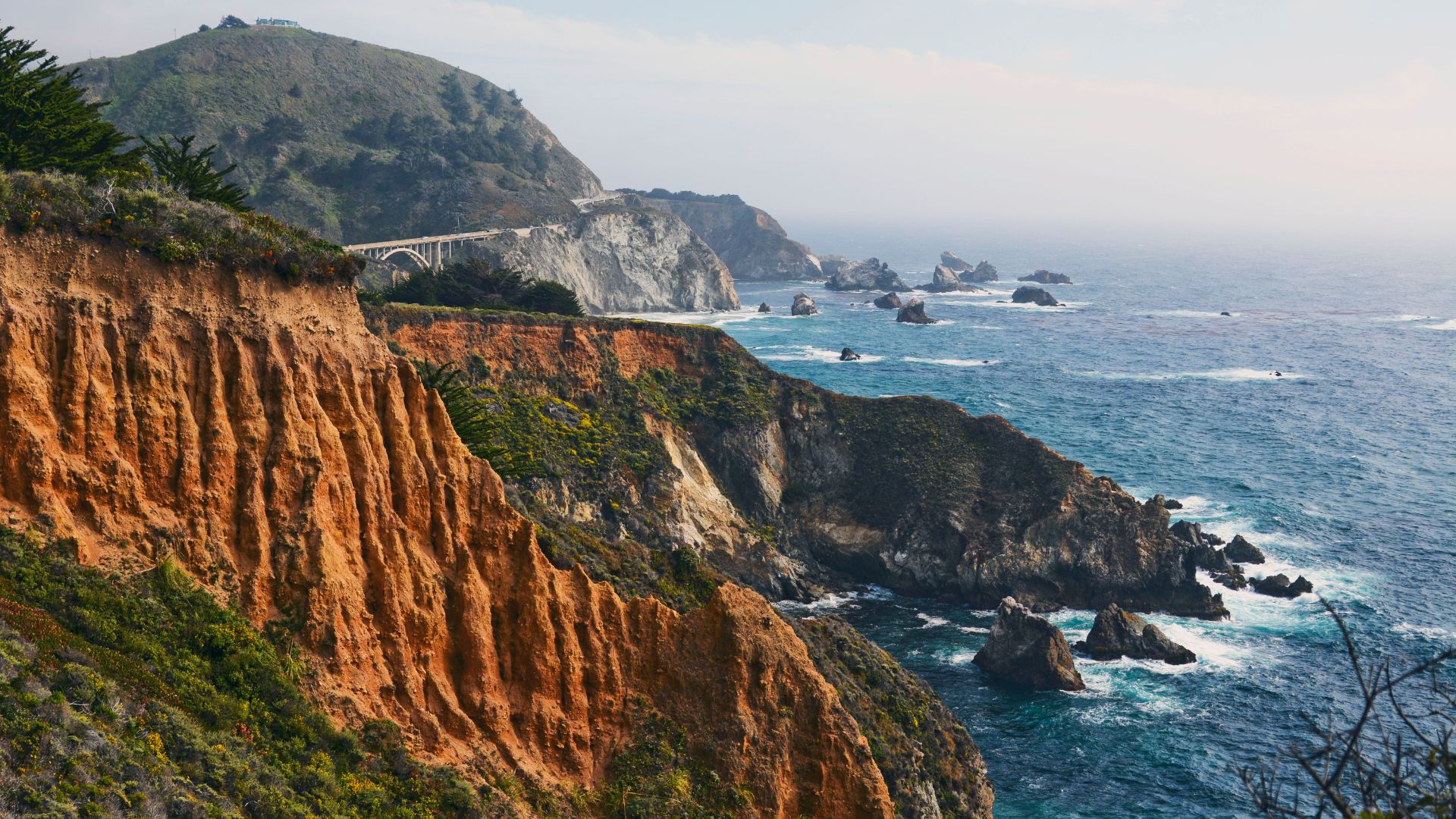 A scenic view of the Big Sur coastline in California, showing rugged cliffs, the Pacific Ocean with crashing waves, and the arch of Bixby Creek Bridge in the distance. 