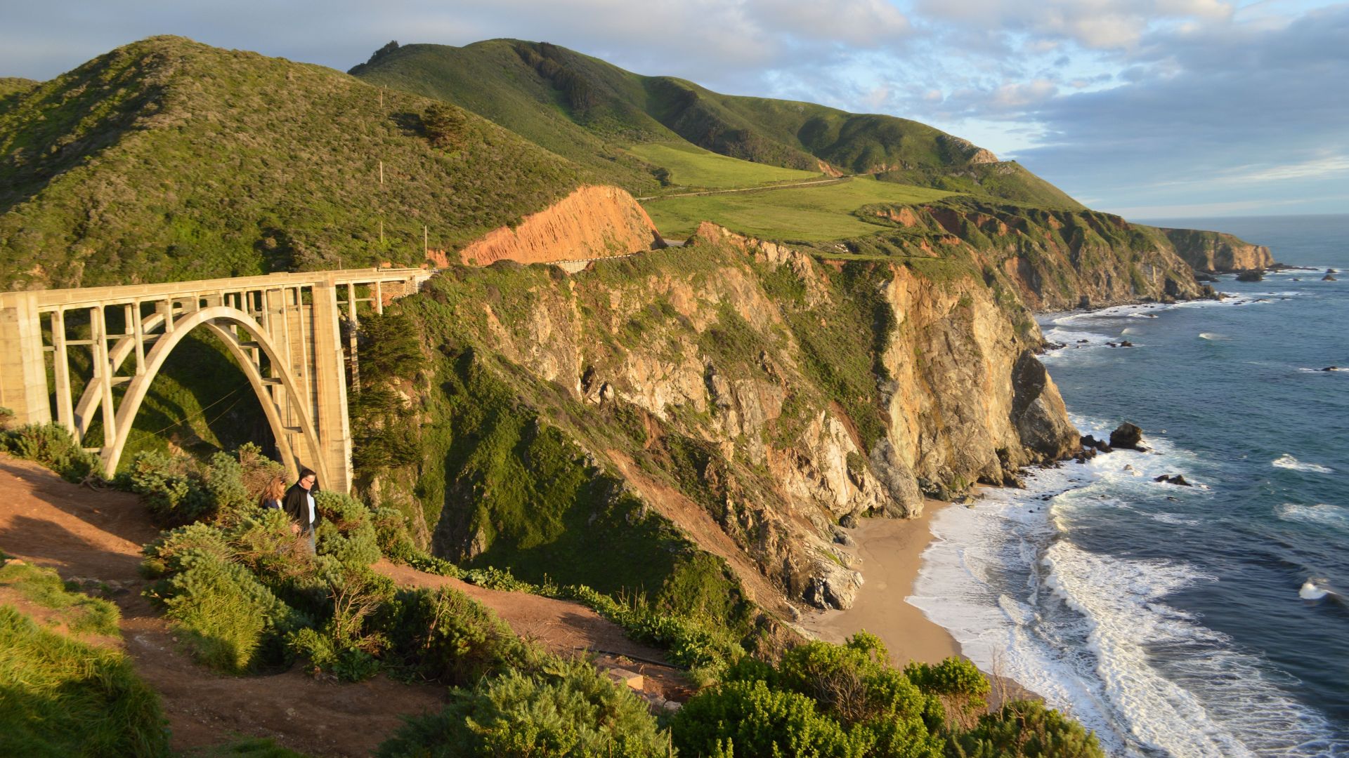 A scenic view of Bixby Creek Bridge, a reinforced concrete open-spandrel arch bridge, spanning a deep canyon along the California coastline, with lush green hills and cliffs meeting the Pacific Ocean on a partly cloudy day. 