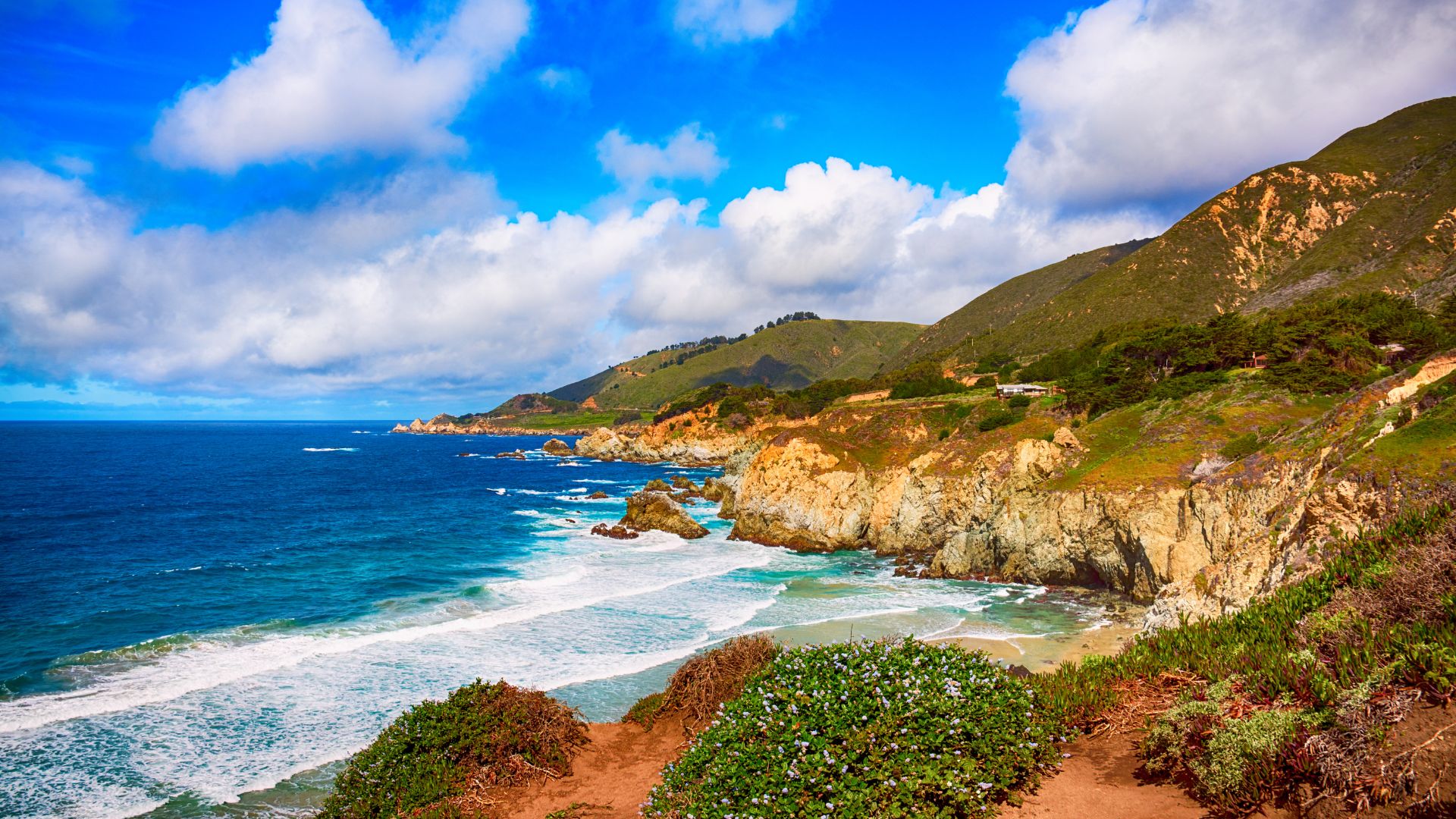 A vibrant coastal landscape of Big Sur, California, featuring rugged cliffs meeting the turquoise ocean with crashing waves, lush green hills, and a clear blue sky with scattered clouds.