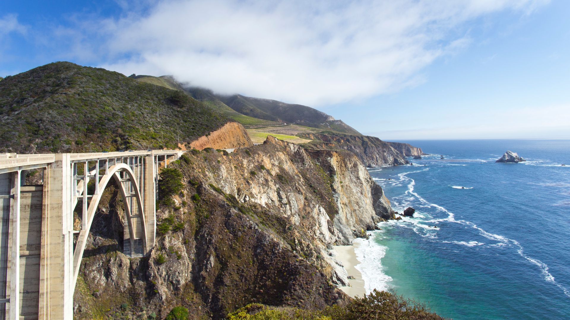 A concrete arch bridge, the Bixby Creek Bridge, spans a dramatic coastal canyon with steep cliffs and a sandy beach below, meeting the Pacific Ocean on a partly cloudy day in Big Sur, California. 