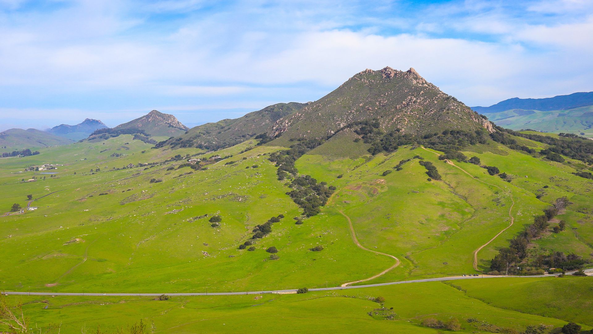 A panoramic view of a prominent, rocky, green-covered peak (Bishop Peak) and surrounding rolling green hills under a blue sky with scattered clouds, with a winding road visible in the foreground.