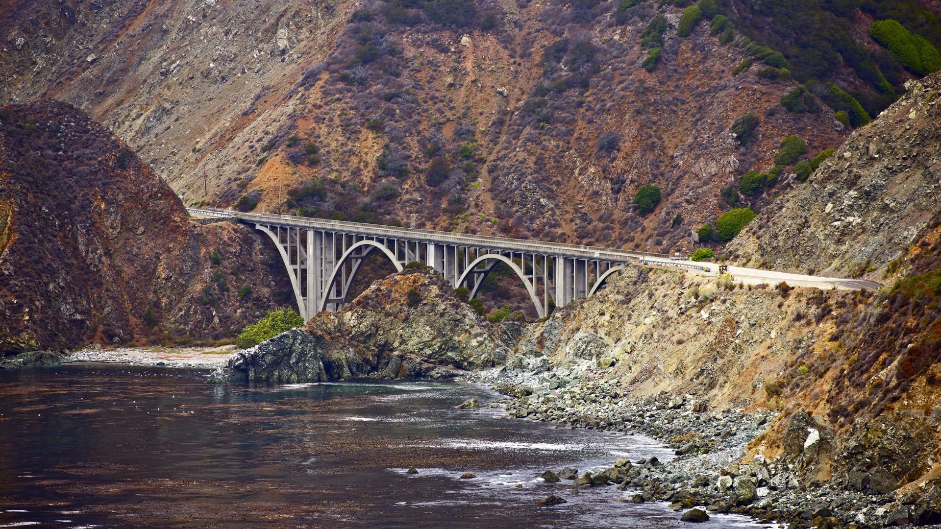 A vibrant coastal landscape of Big Sur, California, featuring rugged cliffs meeting the turquoise ocean with crashing waves, lush green hills, and a clear blue sky with scattered clouds.