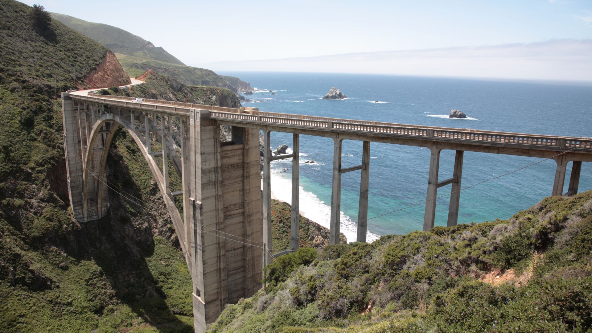 A reinforced concrete open-spandrel arch bridge, the Bixby Creek Bridge, spans a steep canyon with crashing waves of the Pacific Ocean visible below, connecting two lush, green hillsides under a clear blue sky. 