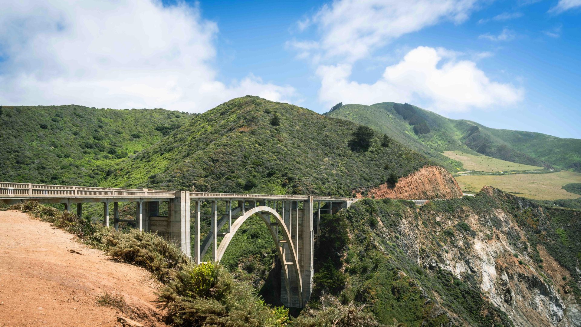 A scenic view of the Bixby Creek Bridge, a large concrete arch bridge, spanning a deep canyon with lush green hills in the background under a blue sky with white clouds. The bridge connects the coastal highway through a dramatic landscape of cliffs and ocean views.