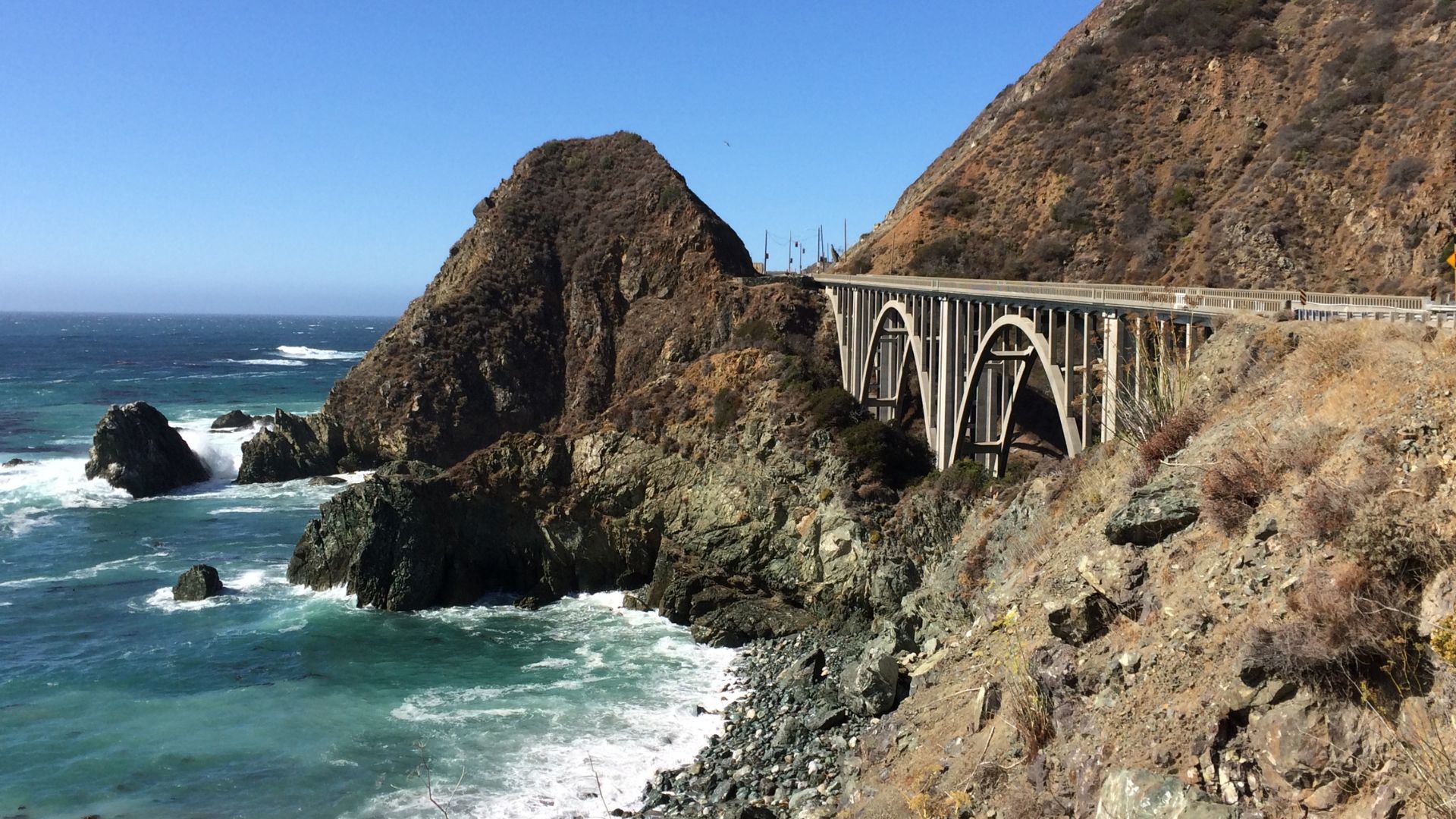 A scenic view of the Big Creek Bridge, a concrete arch bridge, spanning a rugged coastline with crashing waves and steep, rocky cliffs under a clear blue sky in Big Sur, California.