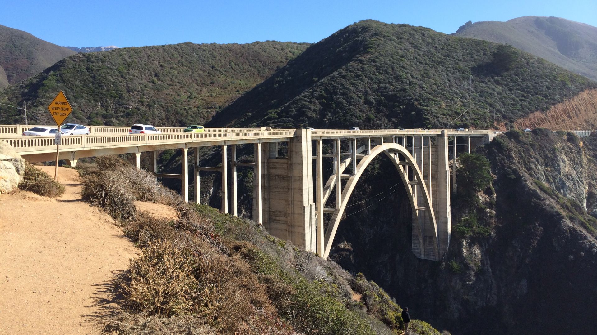 A concrete open-spandrel arch bridge, known as Bixby Creek Bridge, spans a deep canyon with green, brush-covered mountains in the background under a clear sky. Cars are visible on the bridge, and a warning sign is on the left side of the road leading to the bridge. 