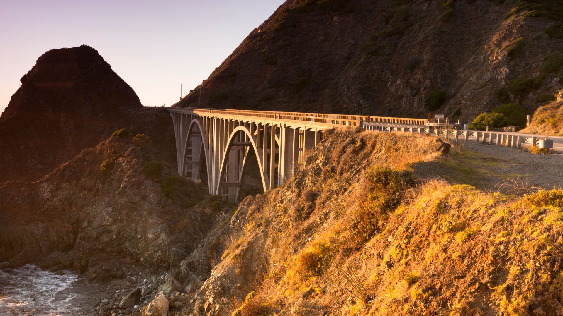An arched concrete bridge spans a rugged coastal canyon with the Pacific Ocean visible below, bathed in golden hour light. The bridge is surrounded by steep, rocky hillsides covered in sparse vegetation.